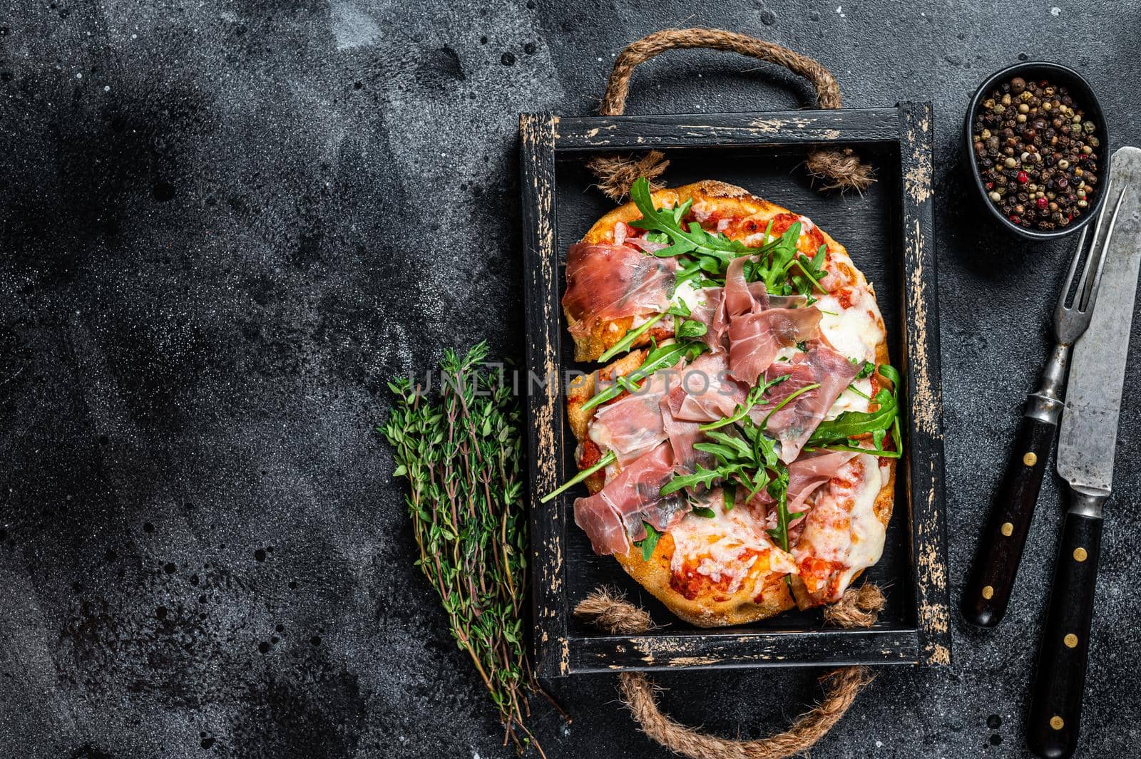 Italian Pizza with prosciutto parma ham, arugula salad and cheese in a rustic wooden tray. Black background. Top view. Copy space by Composter