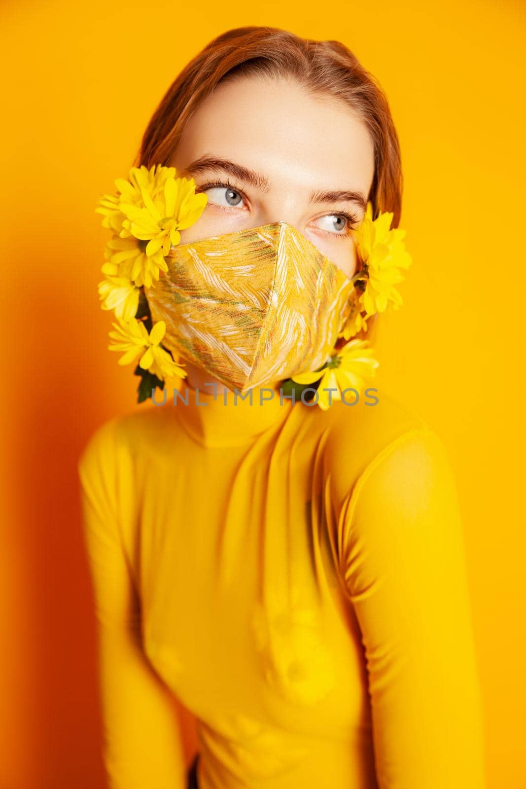 Slim female model in protective mask with fresh yellow flowers standing in transparent outfit on yellow background in studio