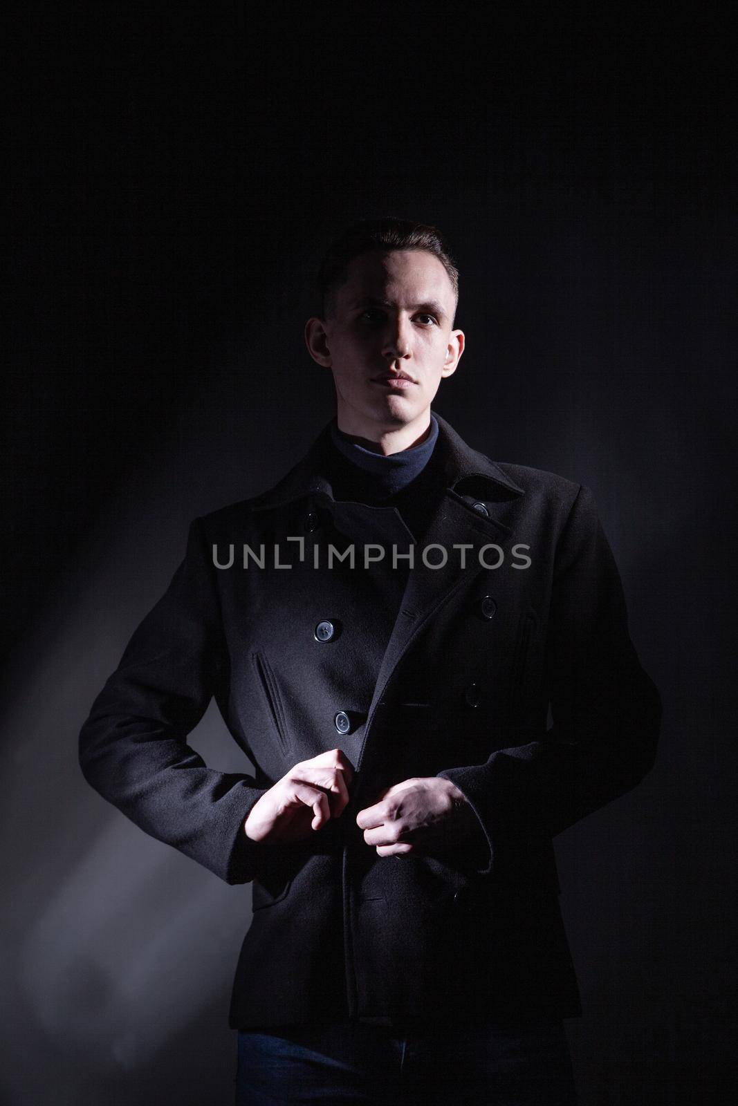 Serious confident young male in black coat looking away while standing under light in dark studio with black wall and shadow