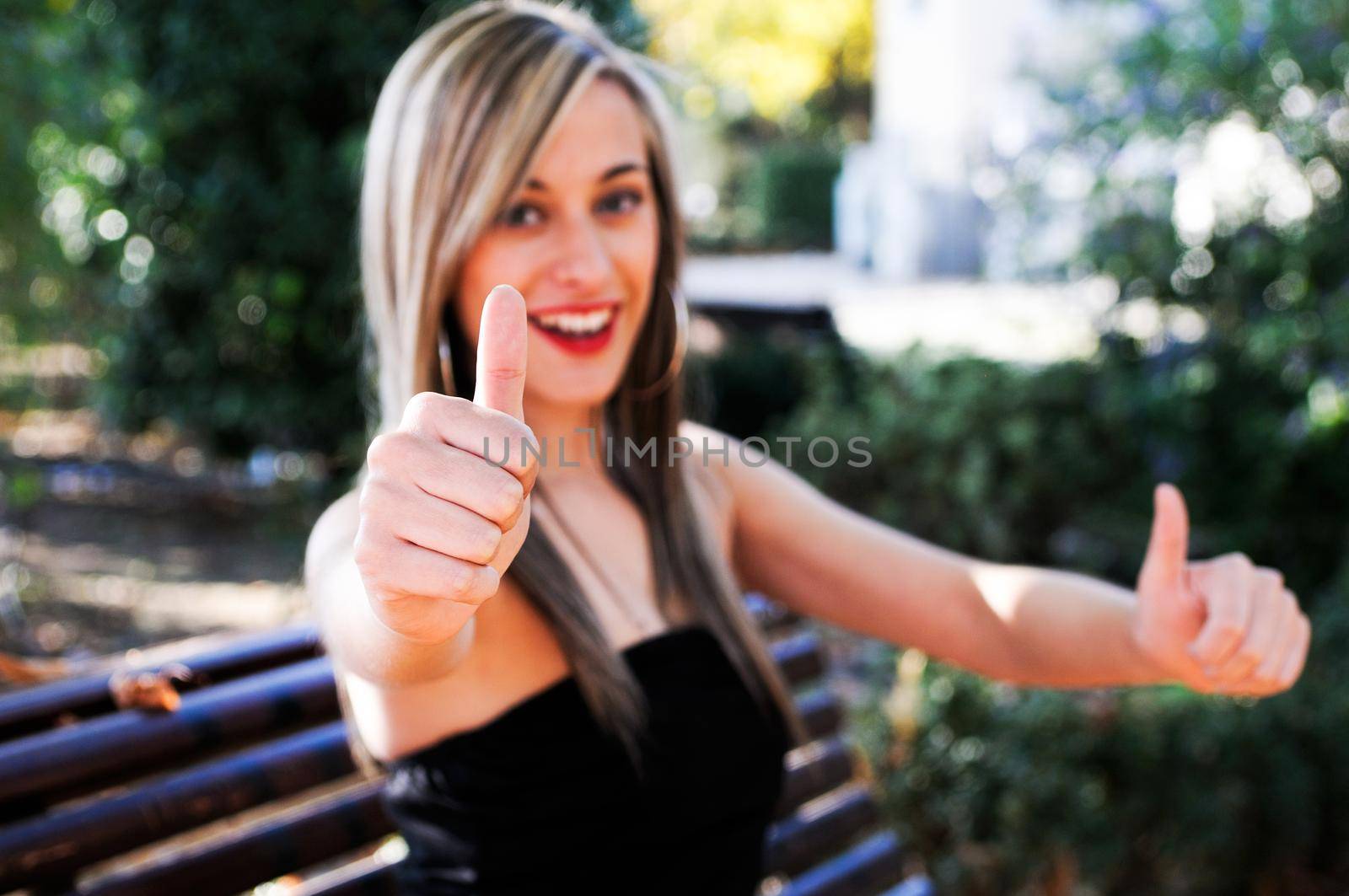 Pretty girl sitting in a bench in the park showing thumb up sign by javiindy