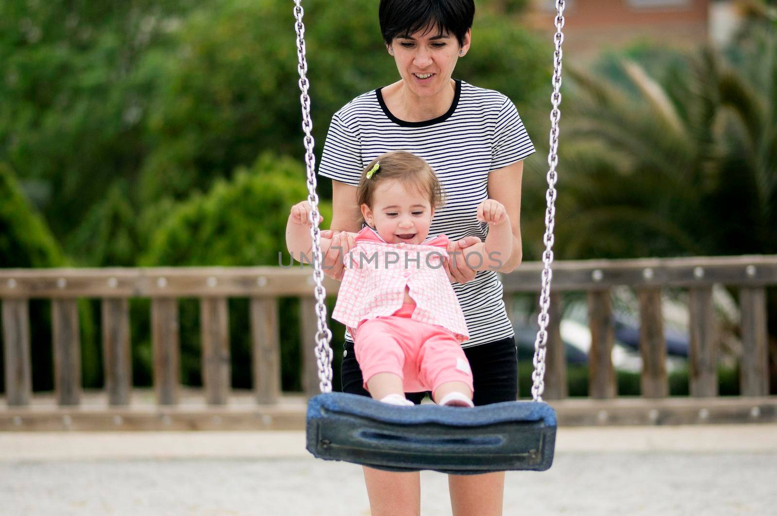 Mother swinging her little daughter on a swing by javiindy