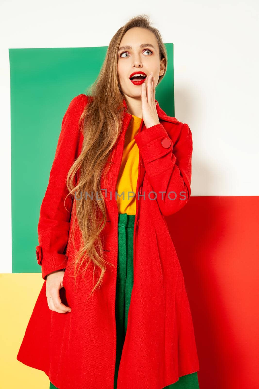 Surprised young fashionable female model in vivid red coat over yellow and green clothes touching cheek and looking away with amazement