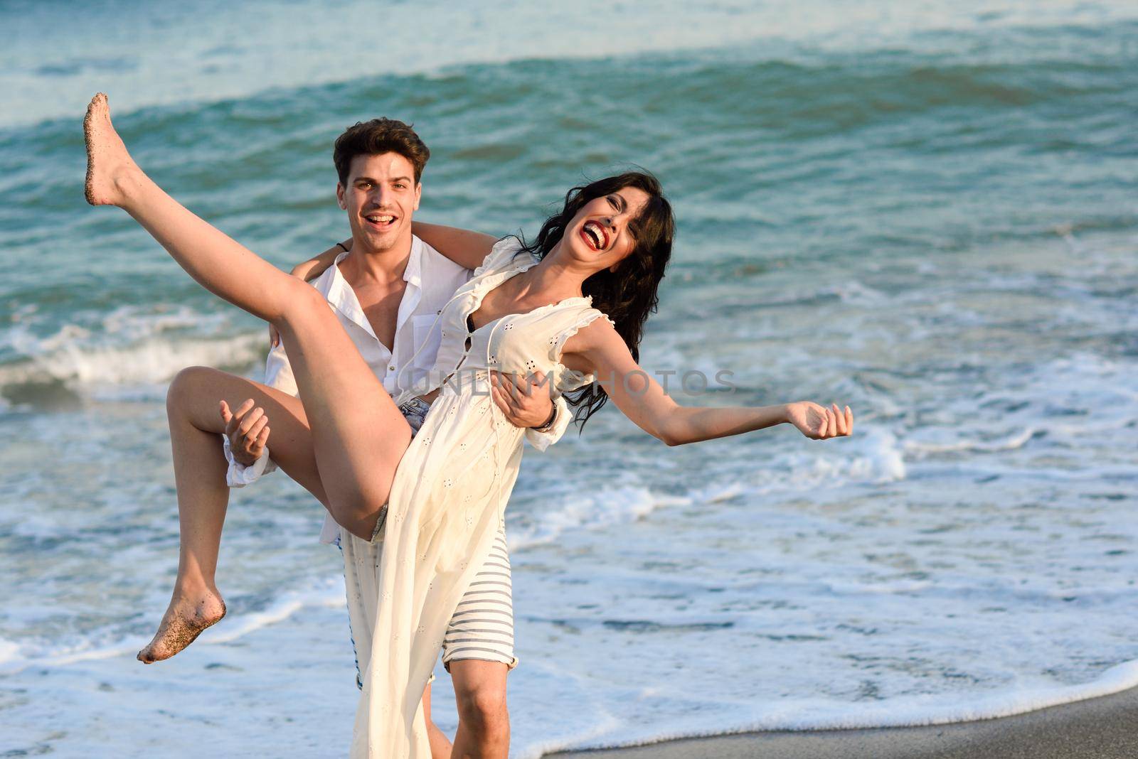 Young happy couple walking in a beautiful beach. Funny Man carrying a woman on his arms. People wearing casual clothes.