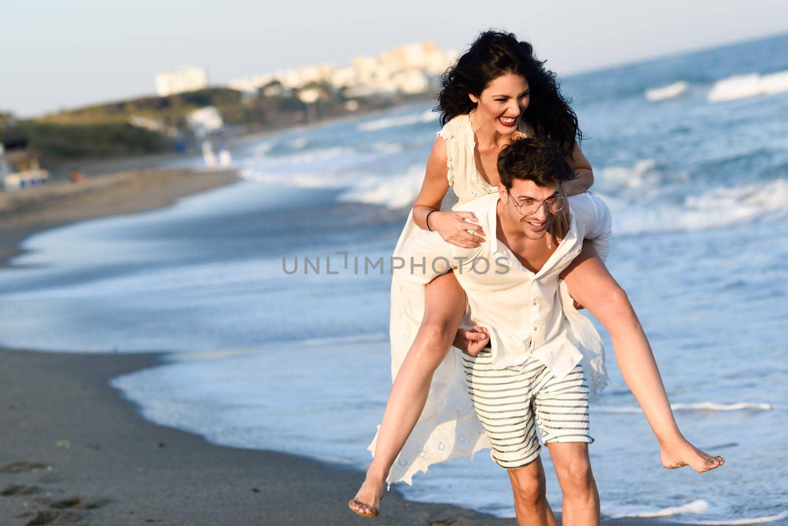 Young happy couple walking in a beautiful beach. Funny Man and woman wearing casual clothes. Male carrying his girlfriend in his arms.