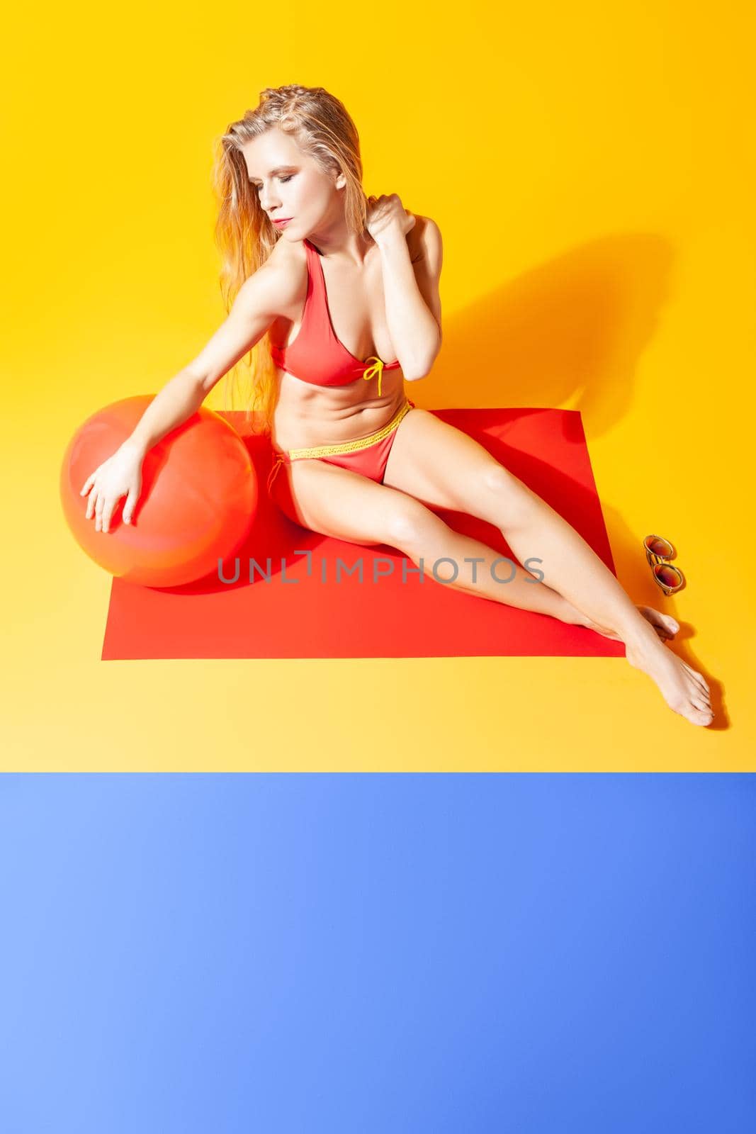 High angle of carefree female in red swimwear sitting with inflatable ball on yellow background in studio while showing concept of summer holidays on beach
