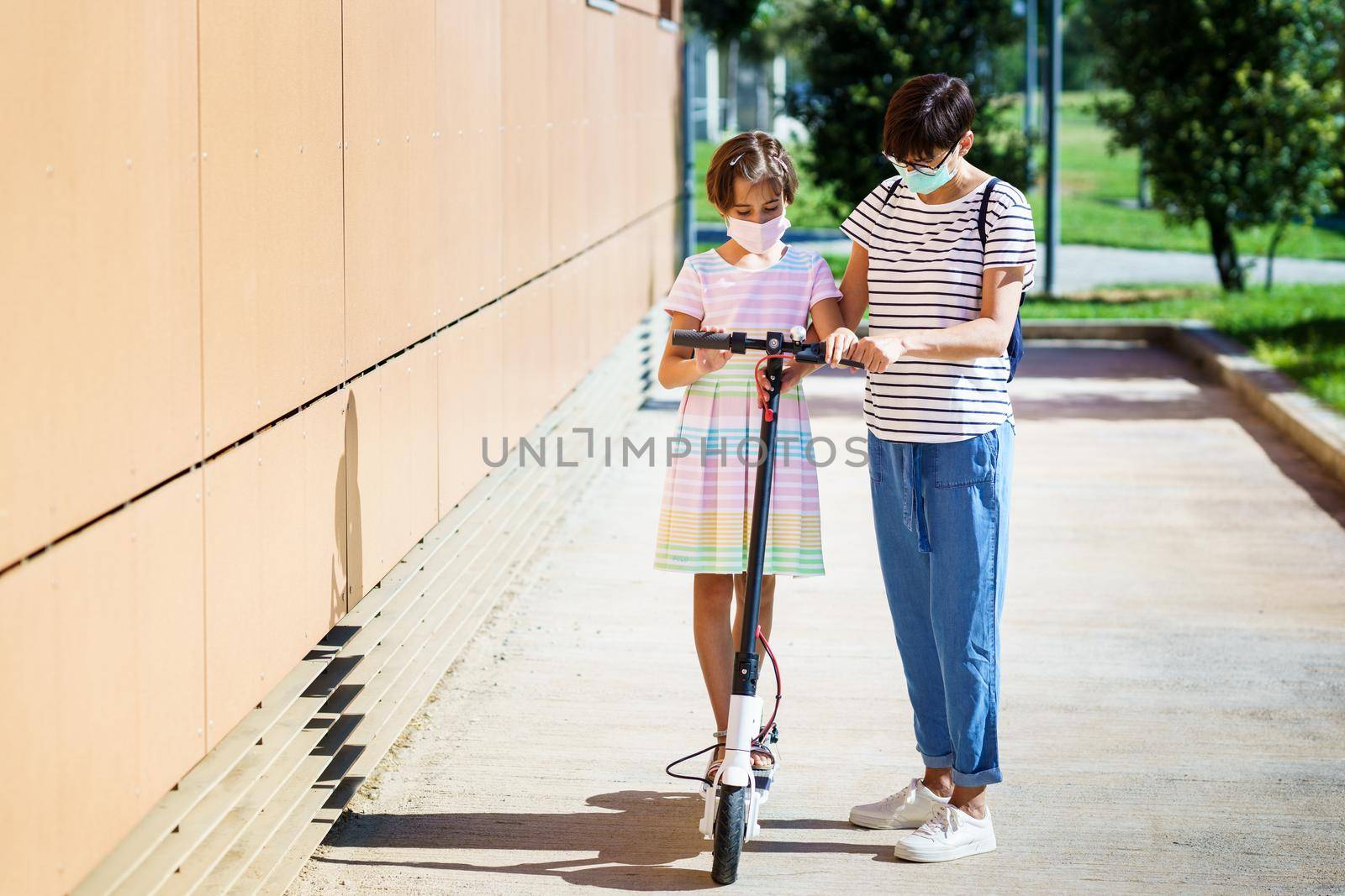 Mother teaching her daughter to ride an electric scooter in urban background wearing surgical masks