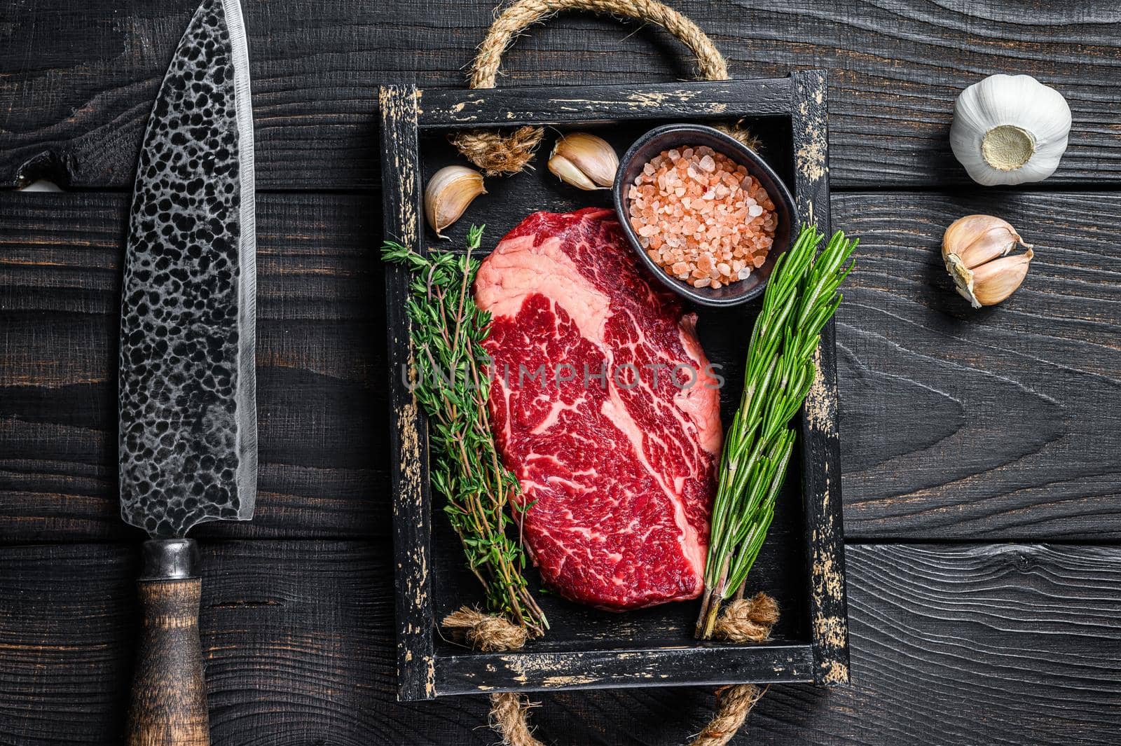 Raw prime rib eye beef meat steak in a butcher wooden tray with herbs. Black wooden background. Top view.