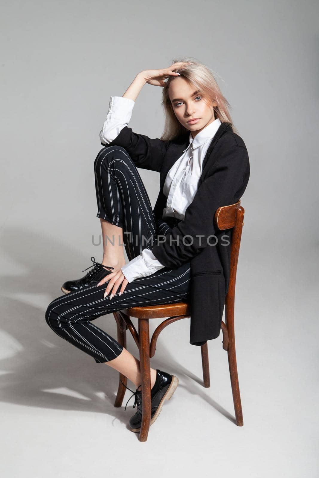Full body young female model in trendy smart casual clothes touching head and leaning back while sitting on chair against gray background