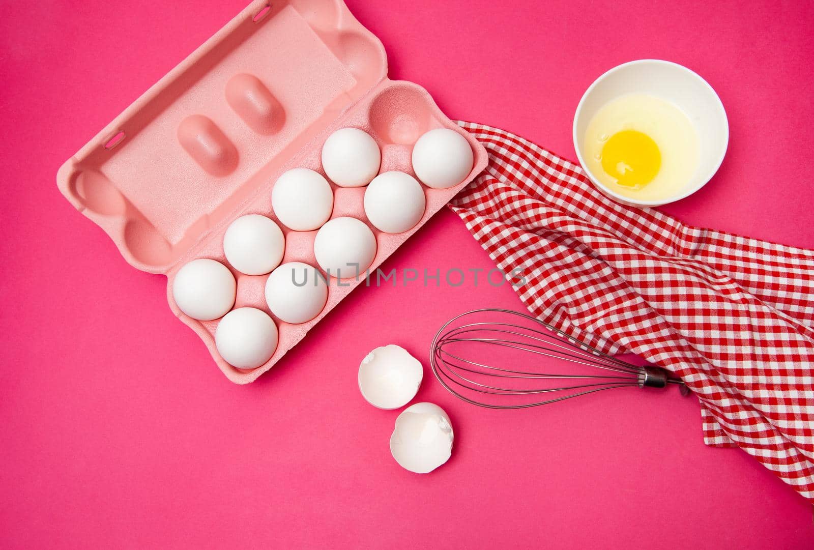 Top view of yolk in bowl placed on pink surface with box of fresh whole eggs near checkered towel and metal hand whisk