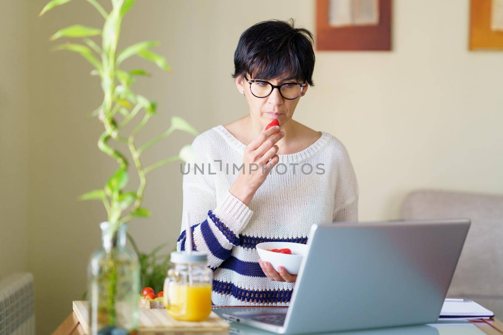 Woman eating strawberries while teleworking from home on her laptop. Female in her 50s