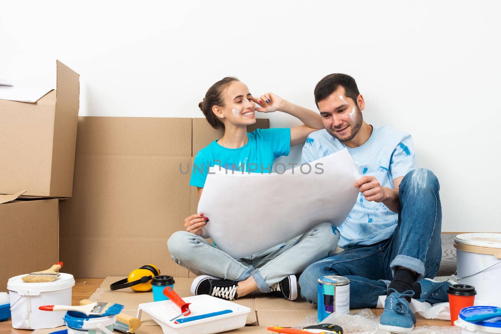 Young man and woman together planning their home renovation. Cardboard boxes, painting tools and materials on floor. House remodeling and interior renovation. People looking at blueprint at home.