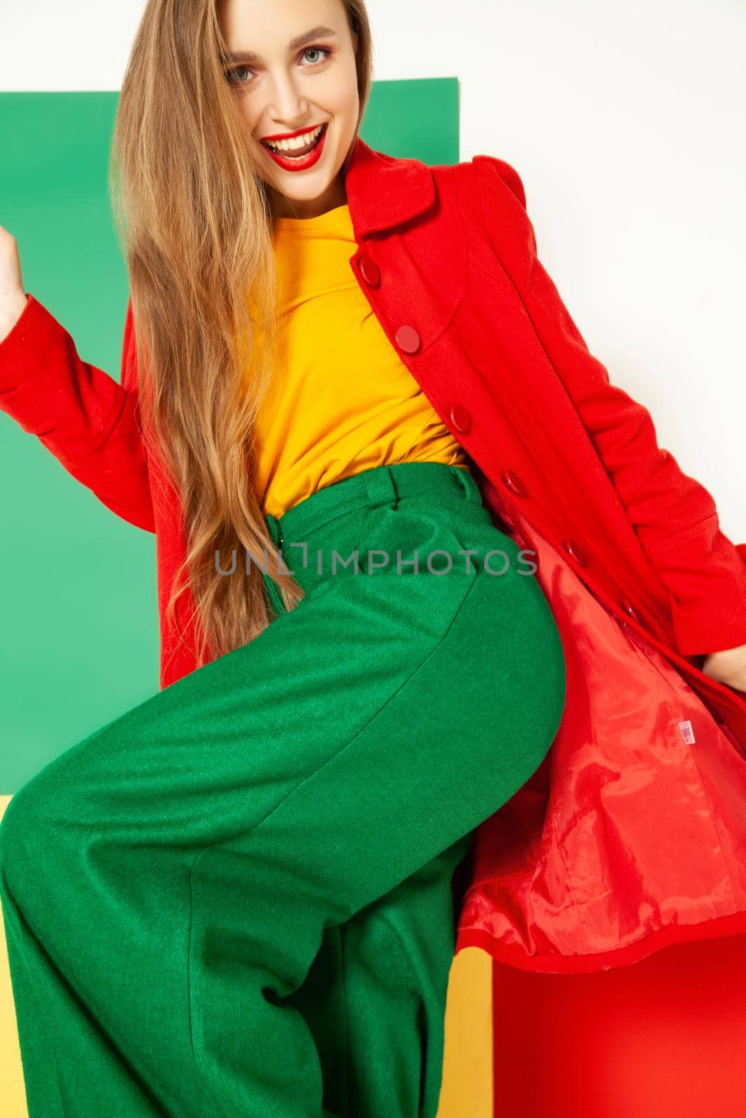 Coquettish young female model in elegant vivid multicolored green red yellow clothes standing against colorful background in studio