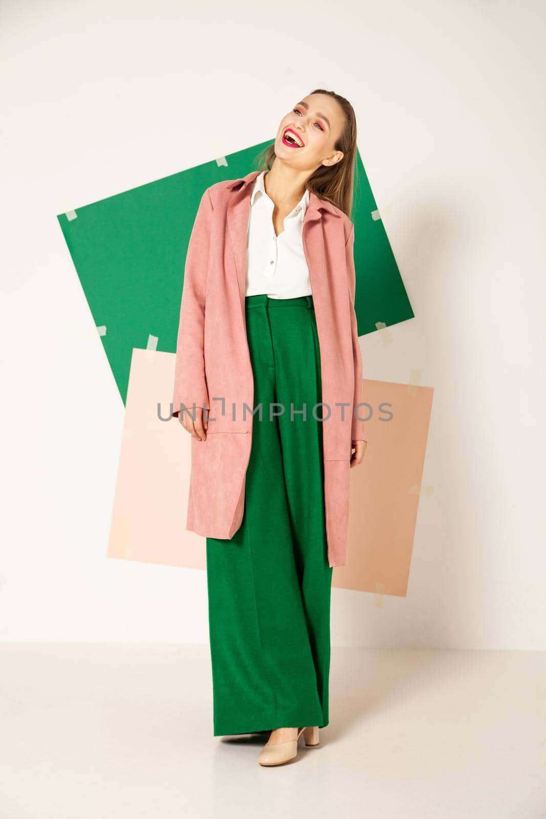 Confident young female model in elegant pastel pink coat and bright green pants representing spring fashion looking away in studio