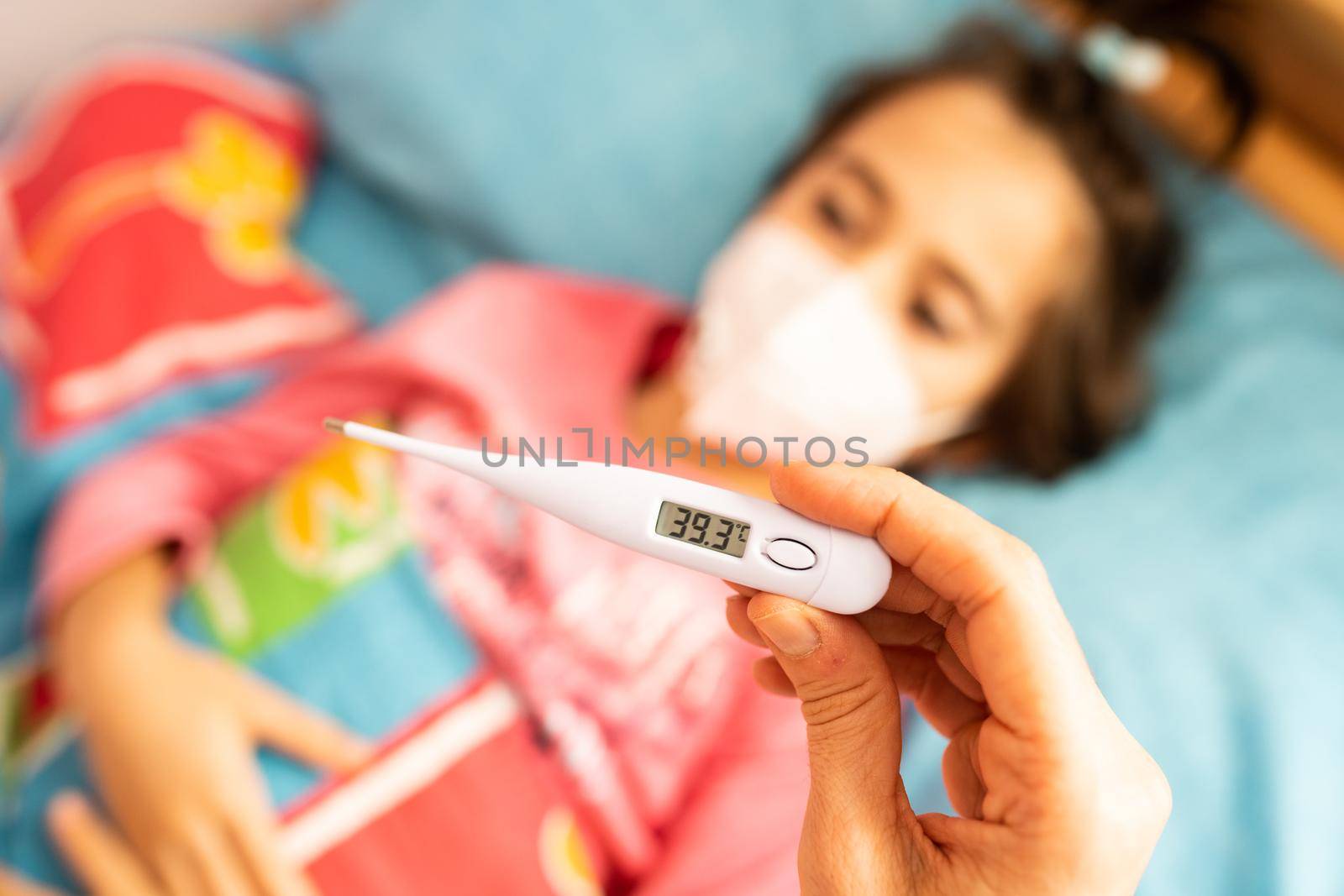 Mother measuring her daughter's temperature, protected by a mask, with a digital thermometer to check for fever