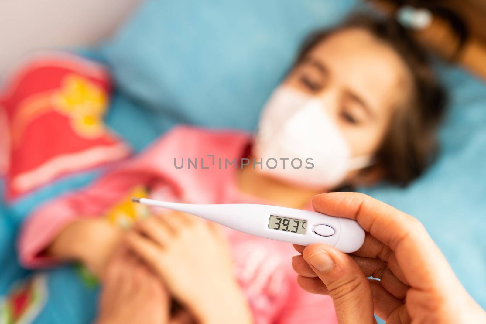 Mother measuring her toddler's temperature, protected by a mask, with a digital thermometer to check for fever