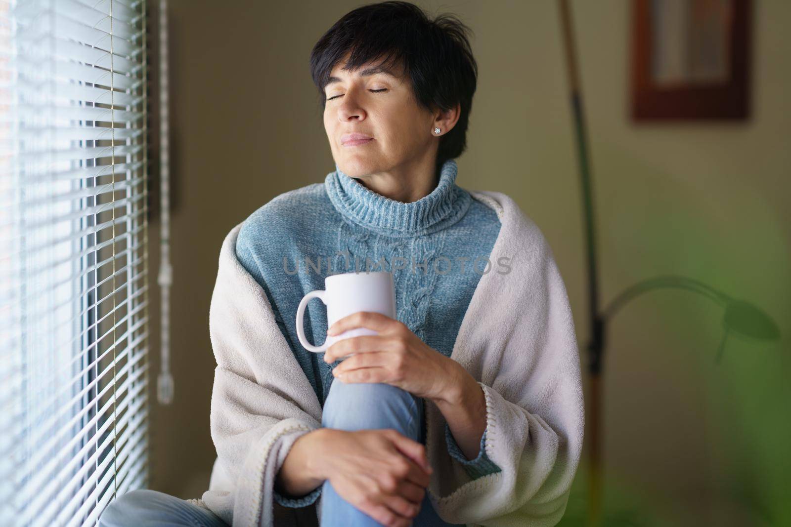 Middle-aged woman with her eyes closed near a window, drinking a mug of coffee. Female in her 50s