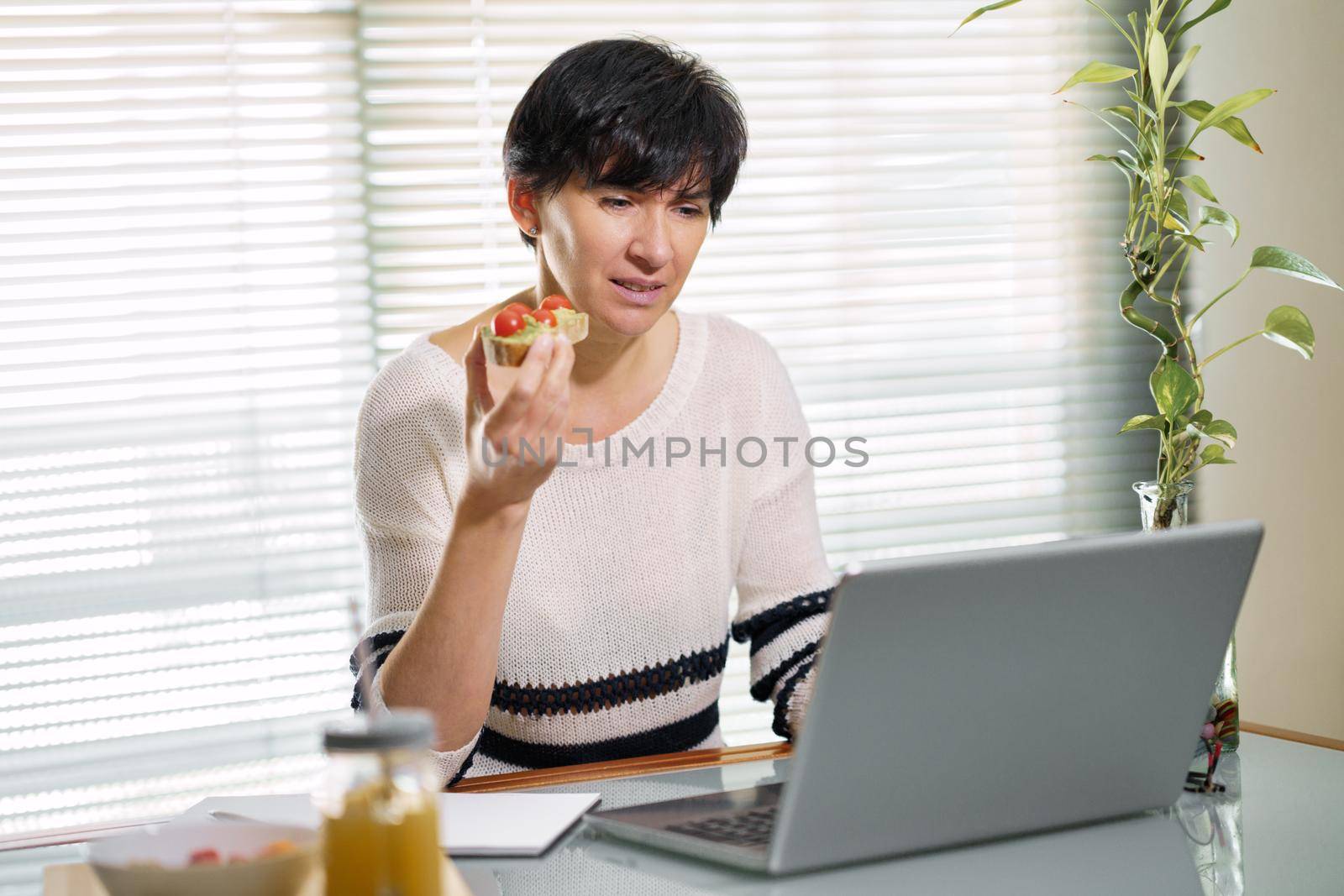 Woman eating some healthy food, while teleworking from home on her laptop. by javiindy