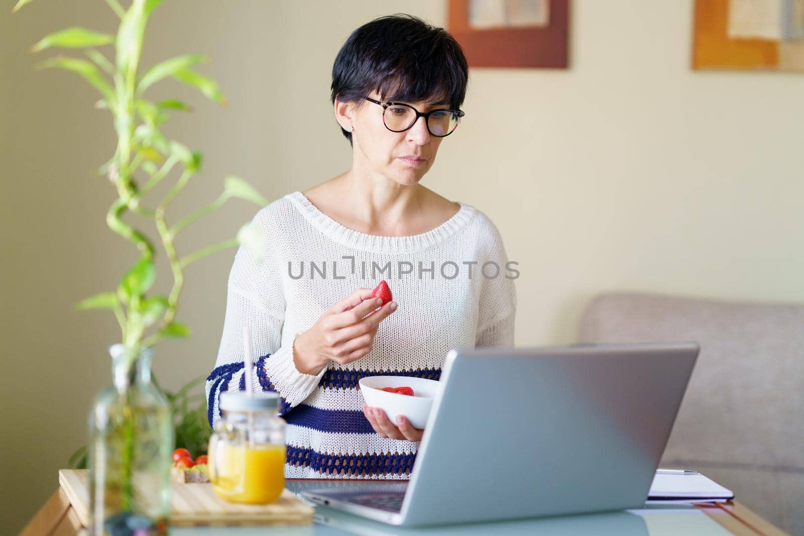 Woman eating strawberries while teleworking from home on her laptop. Female in her 50s