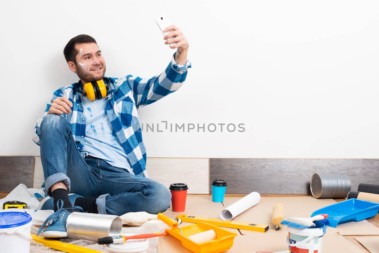 Smiling caucasian man taking selfies photo on mobile phone while sitting near wall. House remodeling and interior renovation. Young bearded guy sitting on floor among construction tools at home.