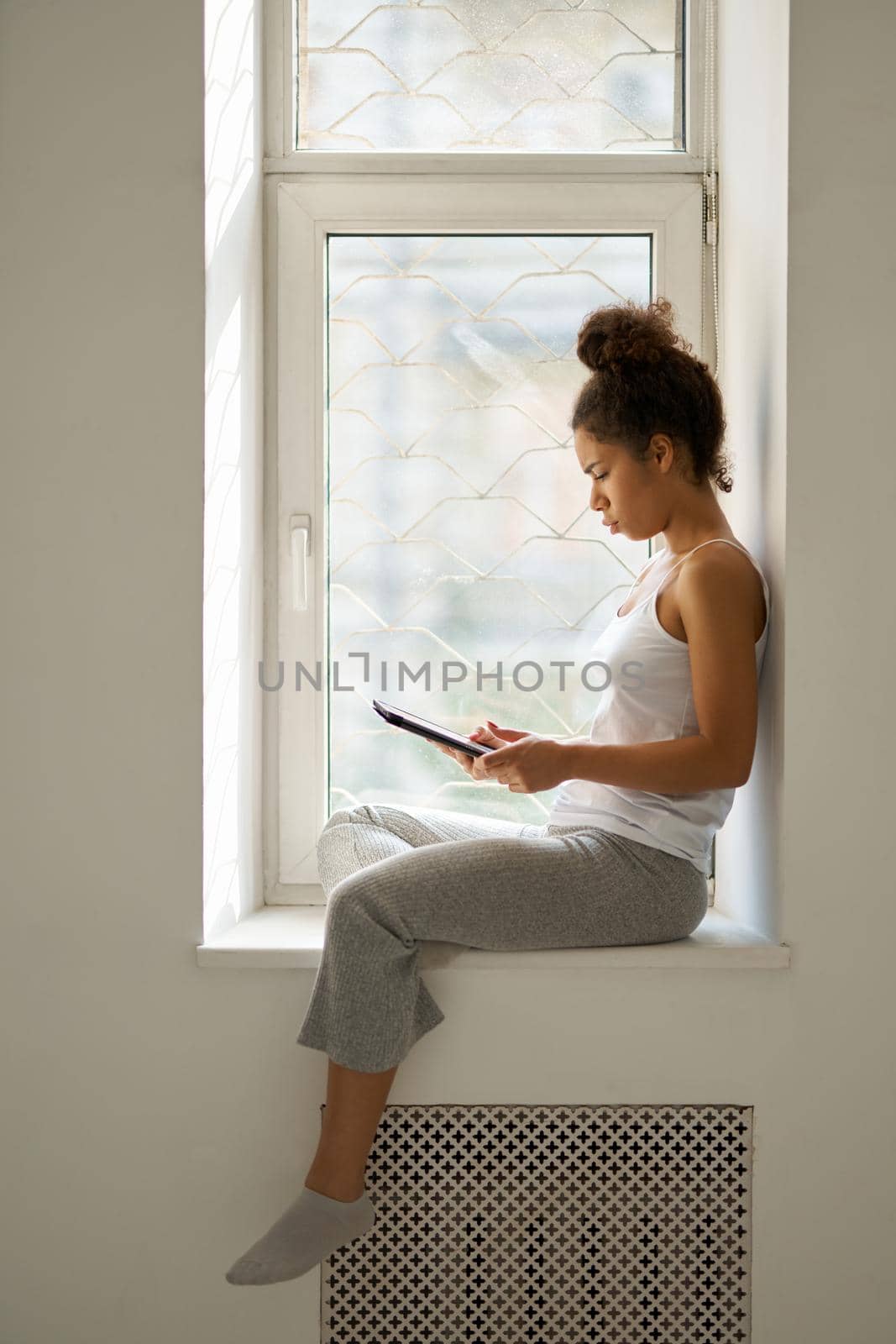 Side view of concentrated young woman reading a book using tablet pc, looking emotional while sitting by the window at home by friendsstock