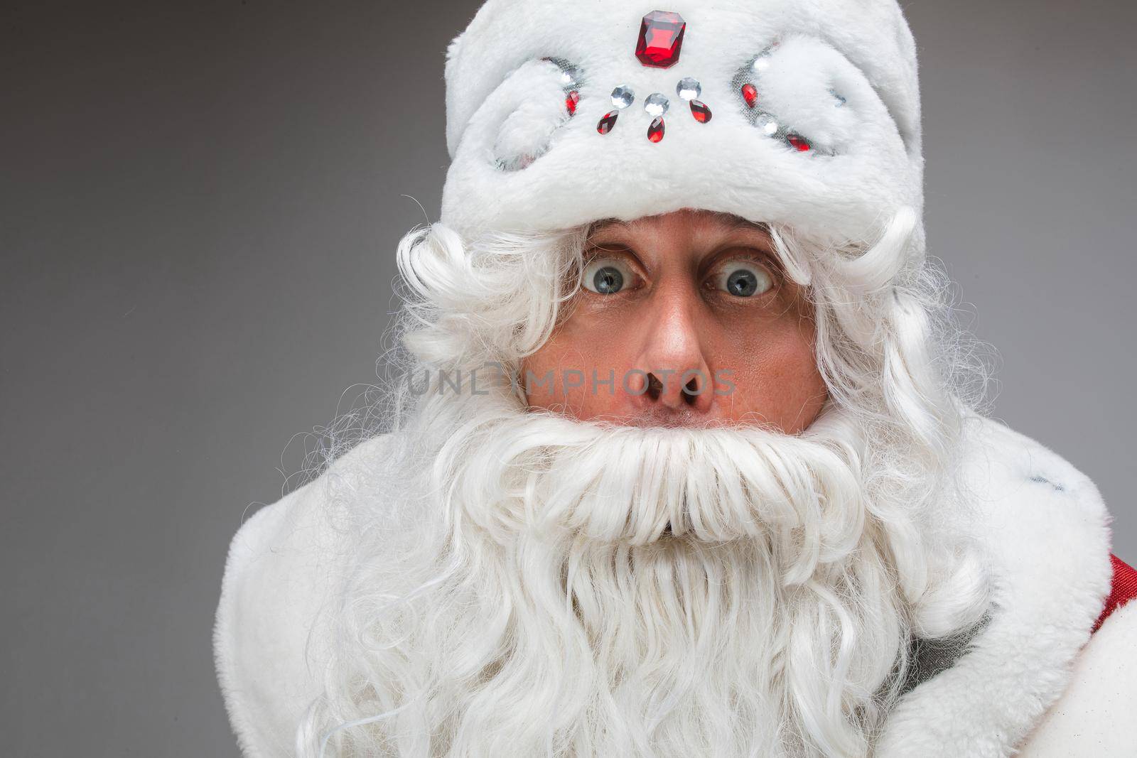 Stock photo of close-up of white-bearded Santa in white fancy hat with rhinestones looking wide-eyed at camera showing astonishment or shock.