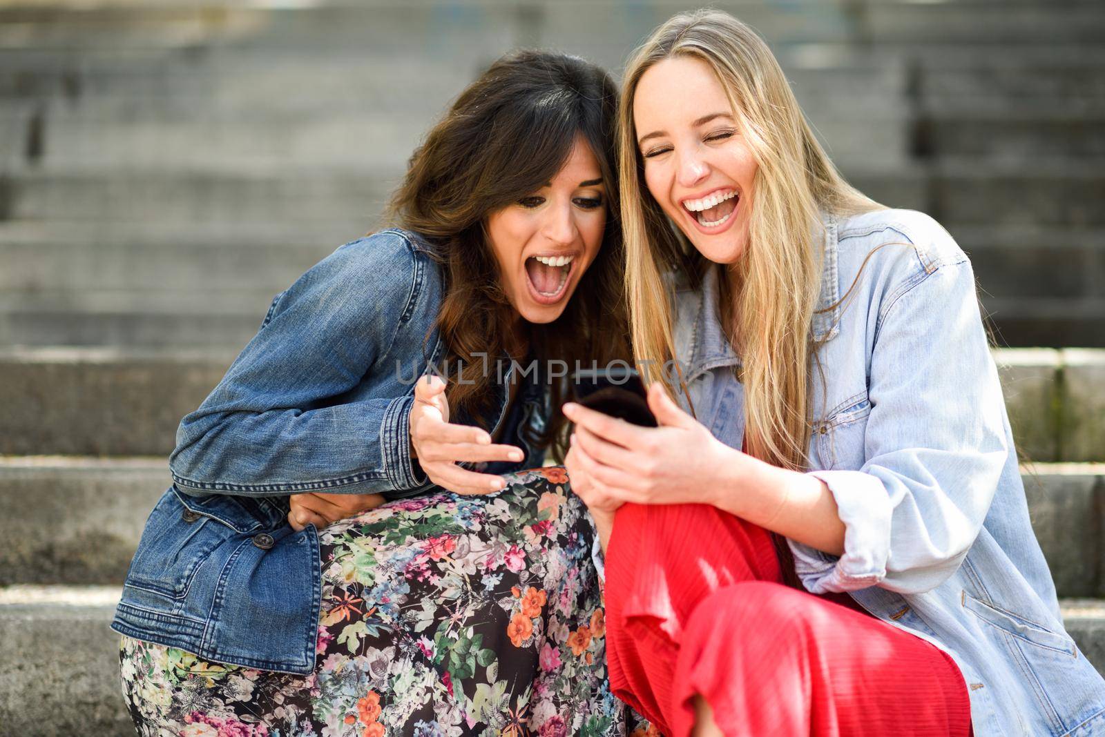 Two young women looking at some funny thing on their smart phone outdoors, sitting on urban steps. Friends girls laughing.