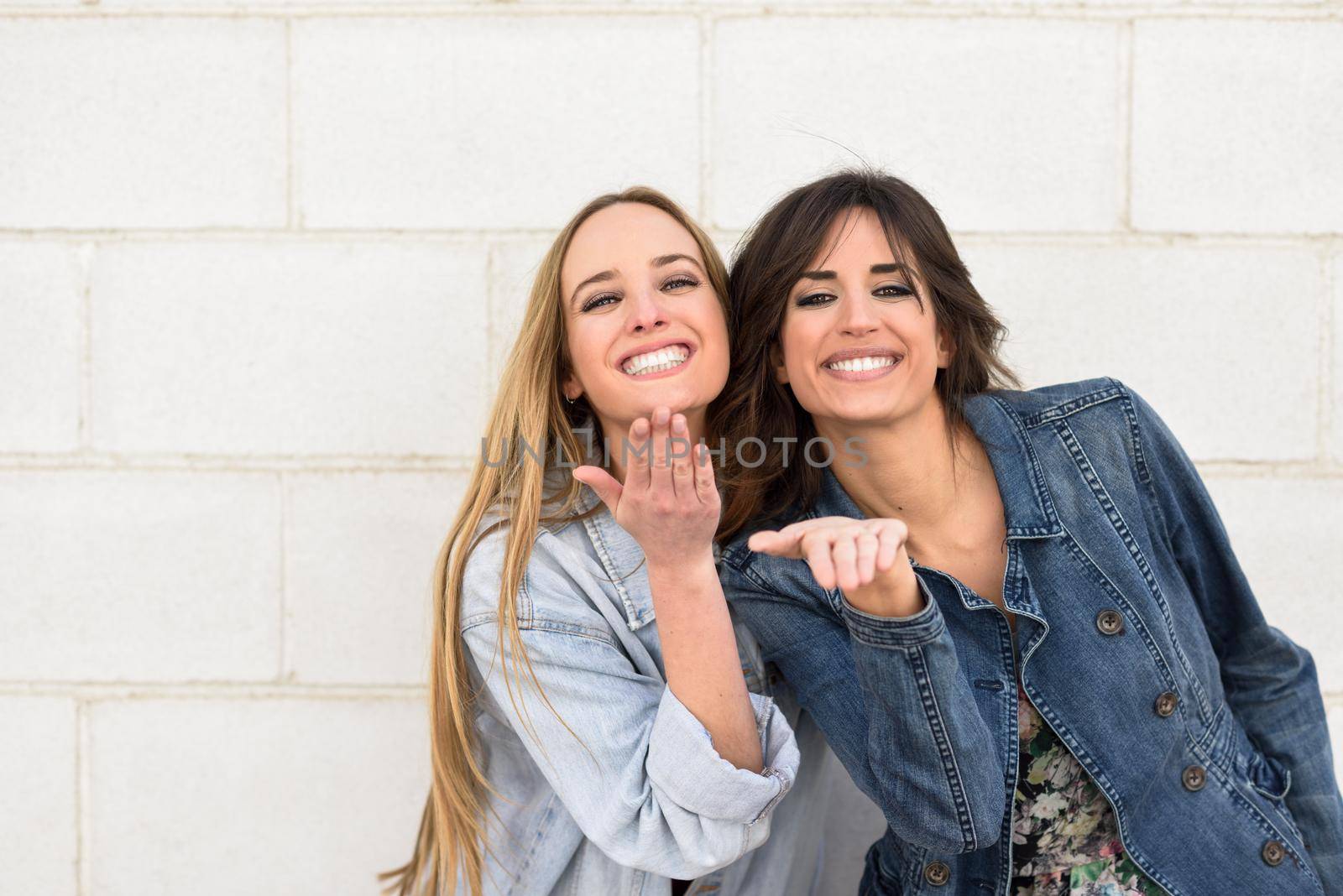 Two beautiful girls blowing a kiss on urban wall outdoors. Young women wearing casual clothes.