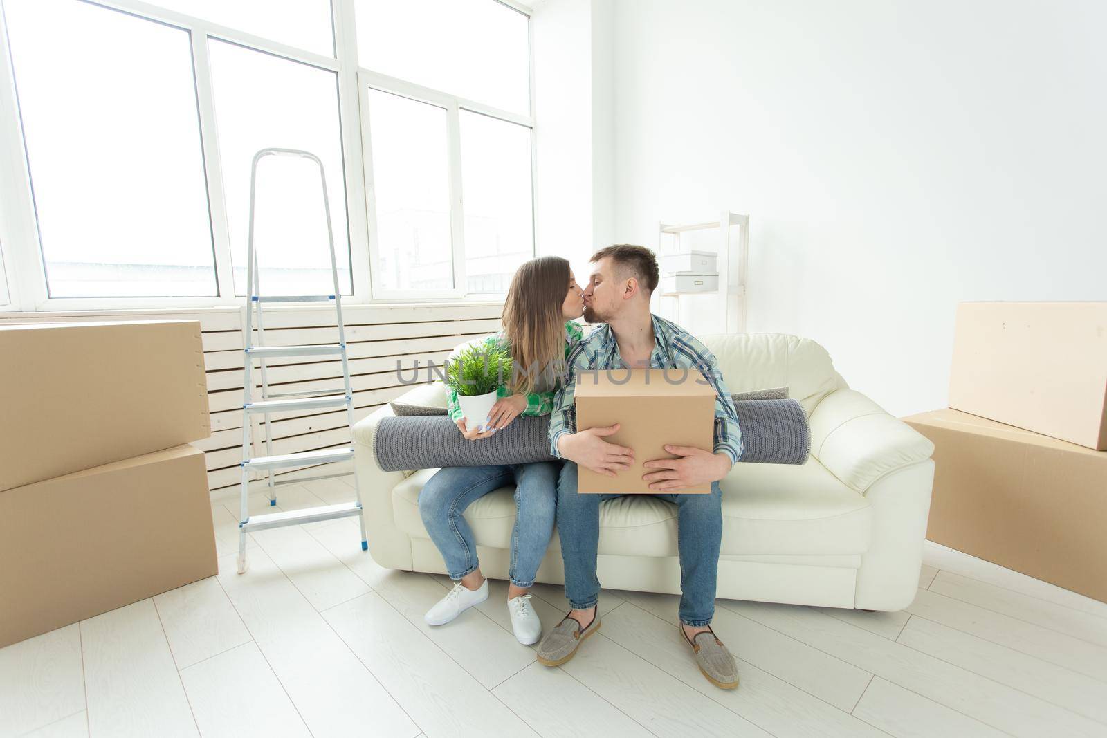 Charming young couple in casual clothes holding things in hands and sitting on sofa kissing to congratulate each other with housewarming. Concept of mortgage and housing affordability for young people