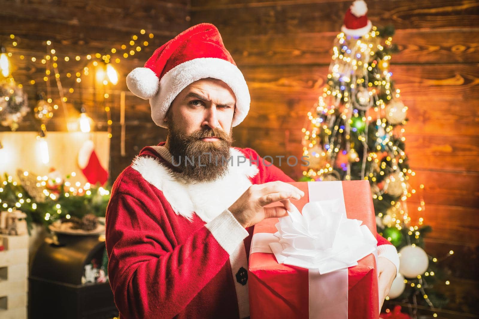 Hipster in red Santa hat holding present. Styling Santa man with a long beard posing on the Christmas wooden background