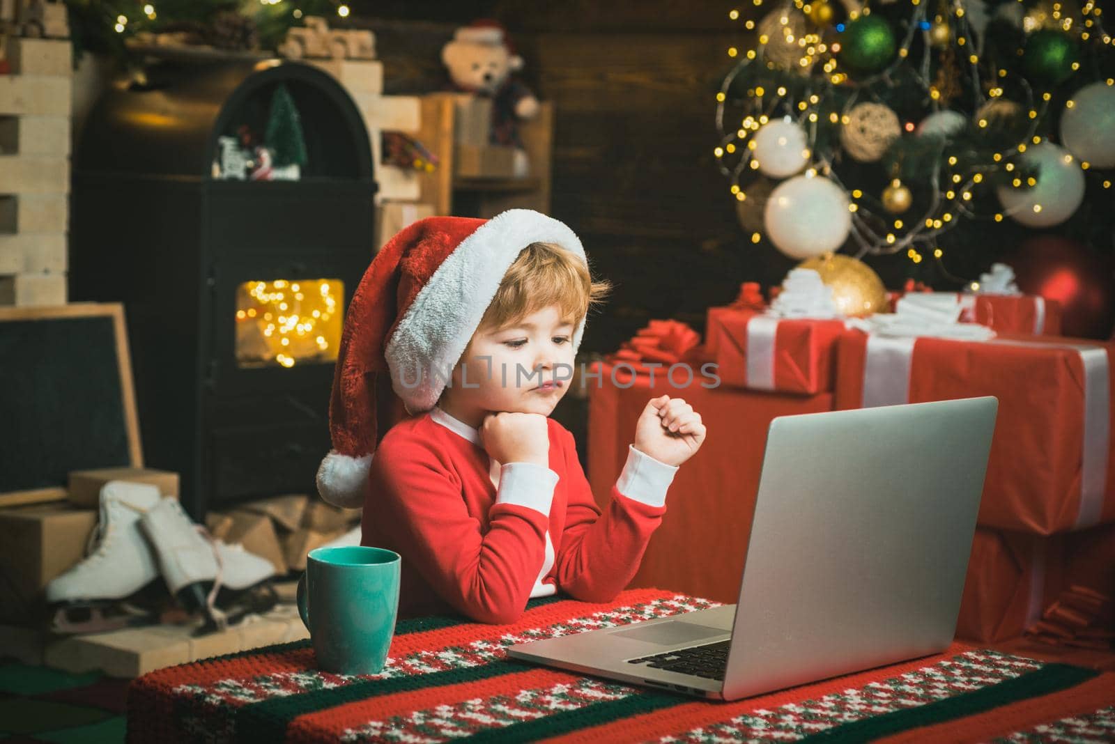 Christmas child boy with computer laptop at home on Christmas background. Little kid is wearing Santa clothes sitting by his laptop. Santa helper using notebook