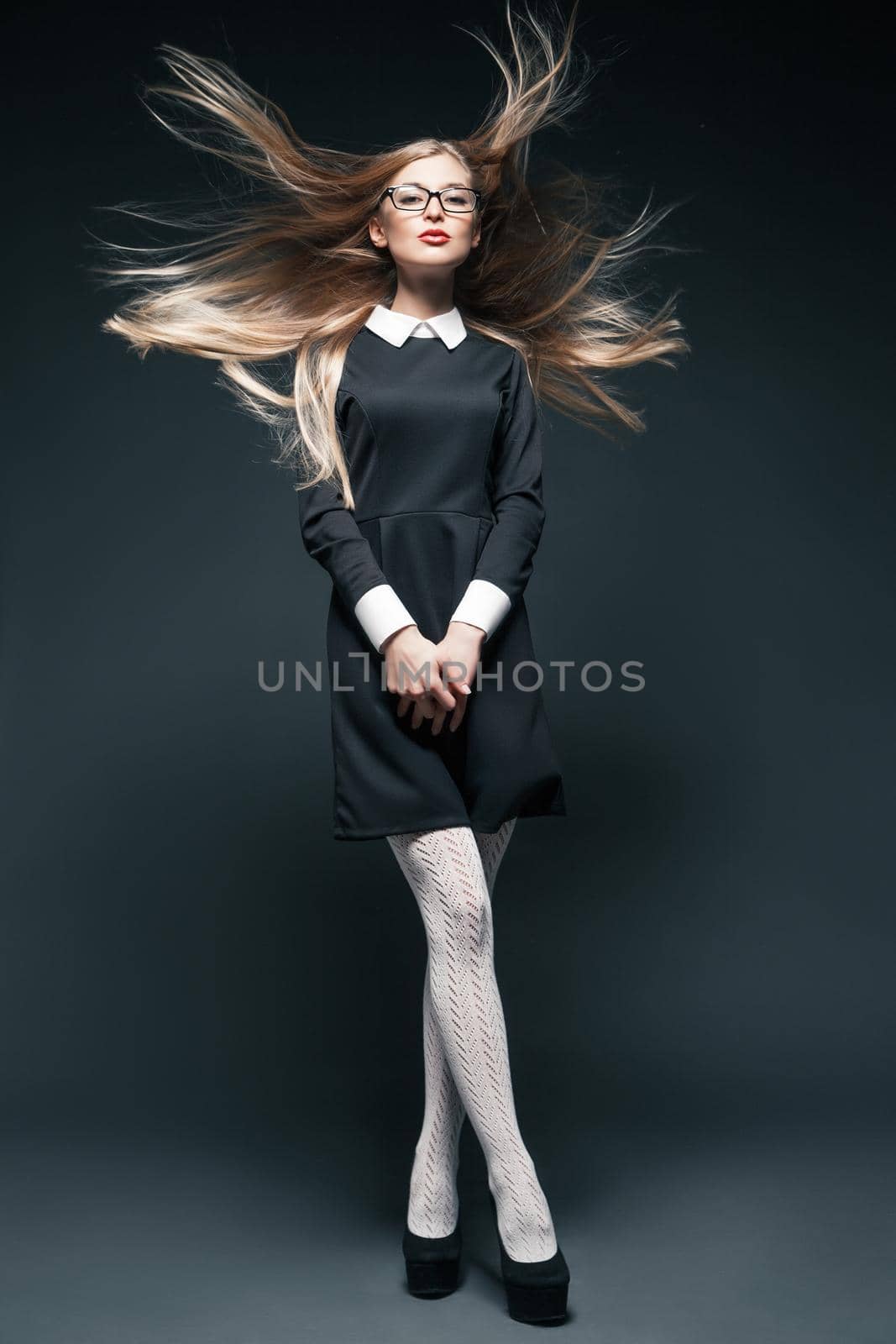 portrait of blonde young beautiful posing woman wearing eyeglasses and black dress with white collar. Her long hair fluttering in wind and legs crossed.