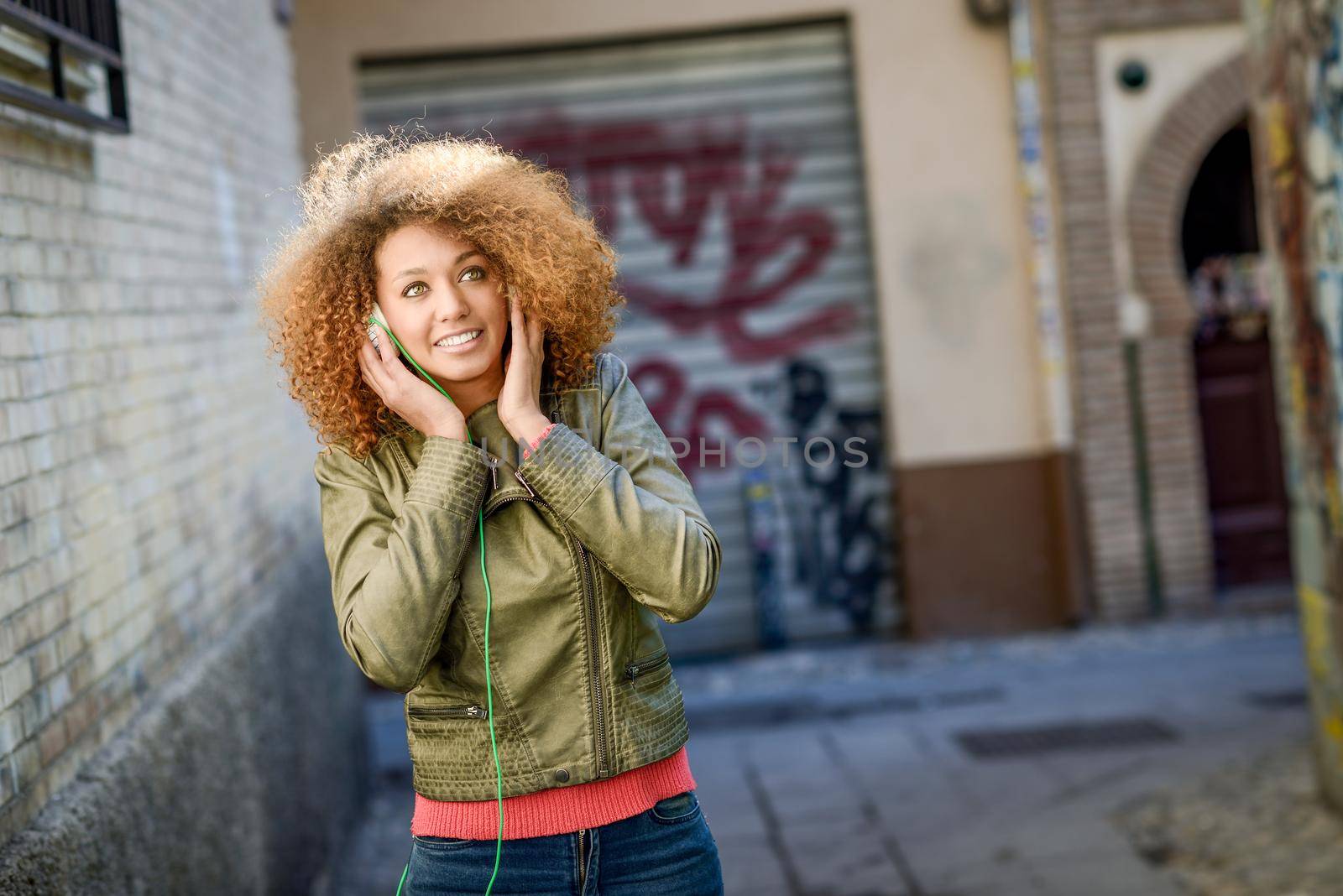 Portrait of young attractive black girl in urban background listening to the music with headphones. Woman wearing leather jacket and blue jeans with afro hairstyle