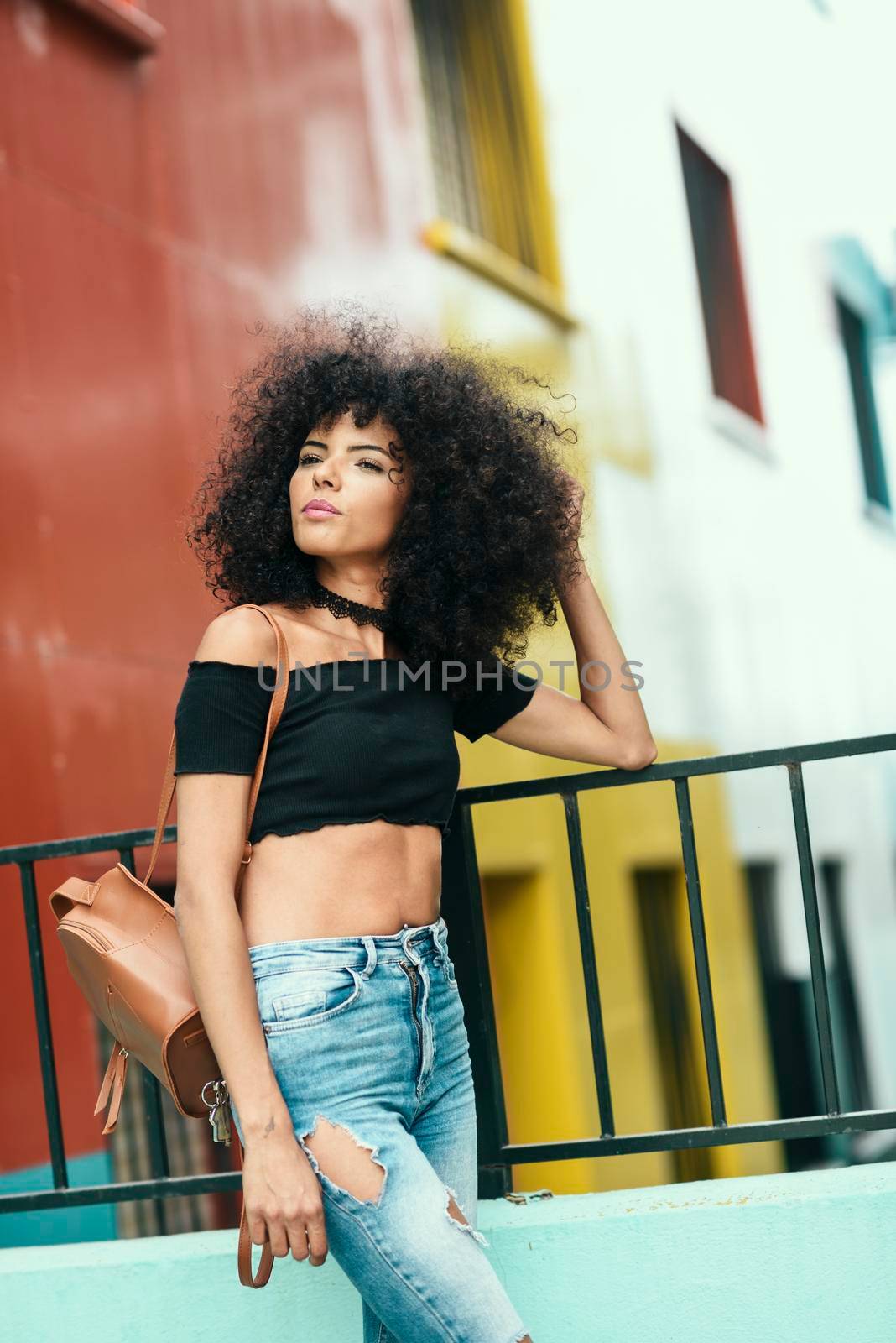 Young mixed woman with curly hair near a modern colorful building. Female wearing casual clothes outdoors. Lifestyle concept