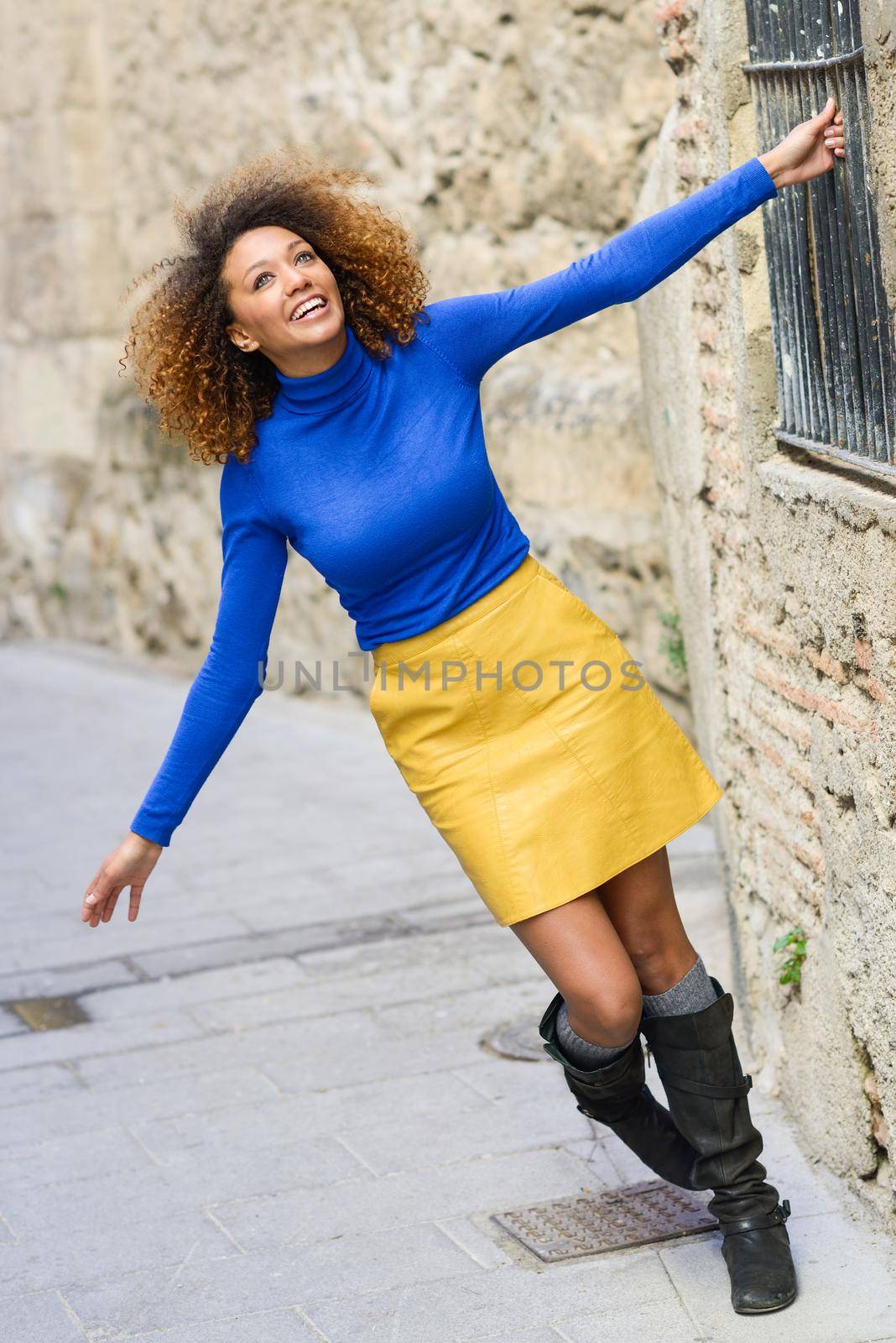 Funny young African American woman, model of fashion, smiling with afro hairstyle and green eyes wearing blue sweater and yellow skirt in urban background