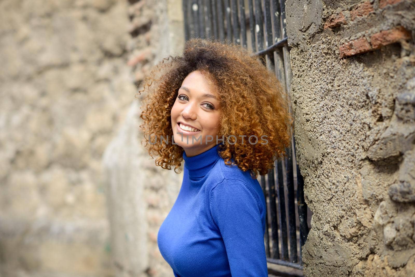 Beautiful young African American woman, model of fashion, smiling with afro hairstyle and green eyes wearing blue sweater in urban background