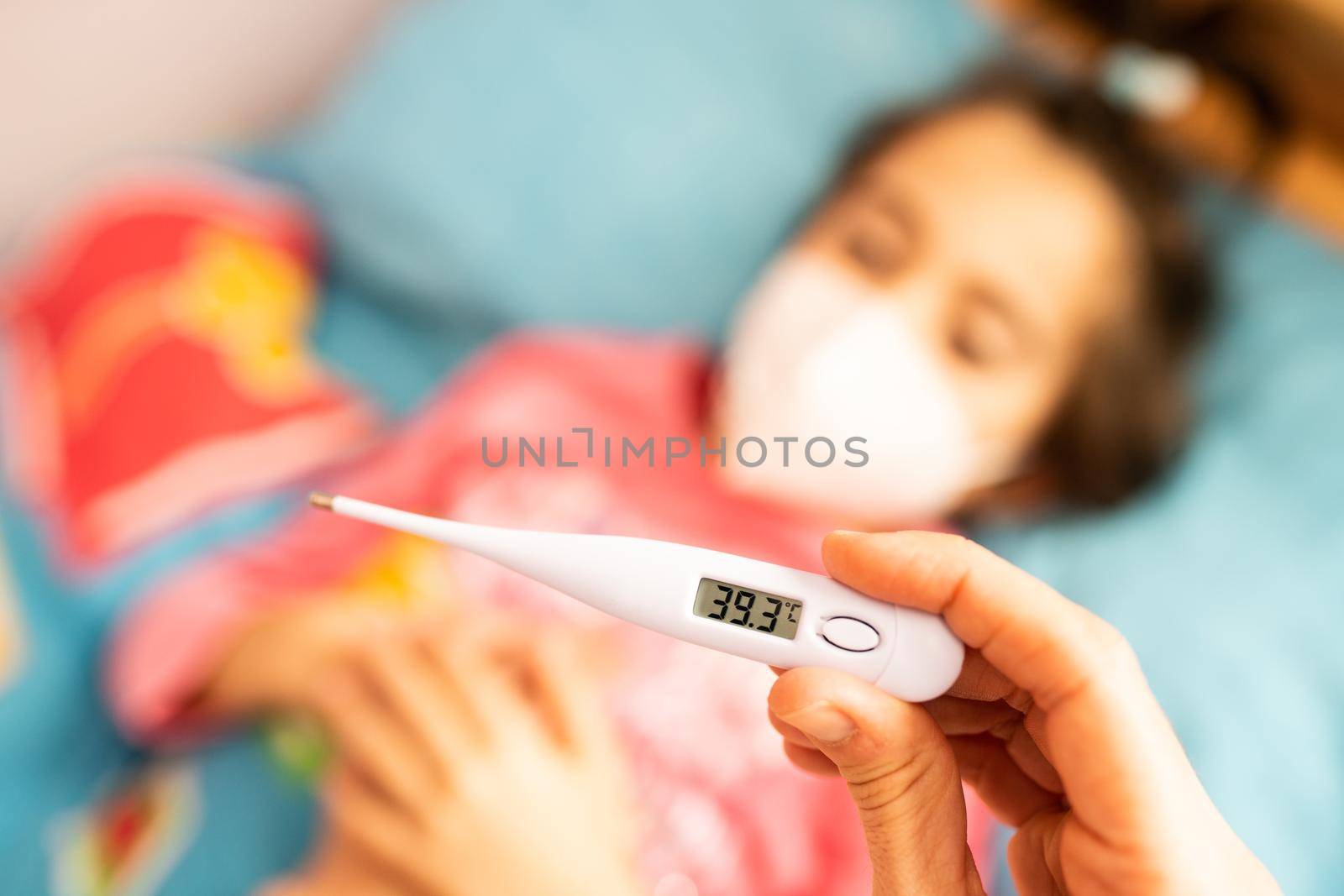 Mother measuring her daughter's temperature, protected by a mask, with a digital thermometer to check for fever