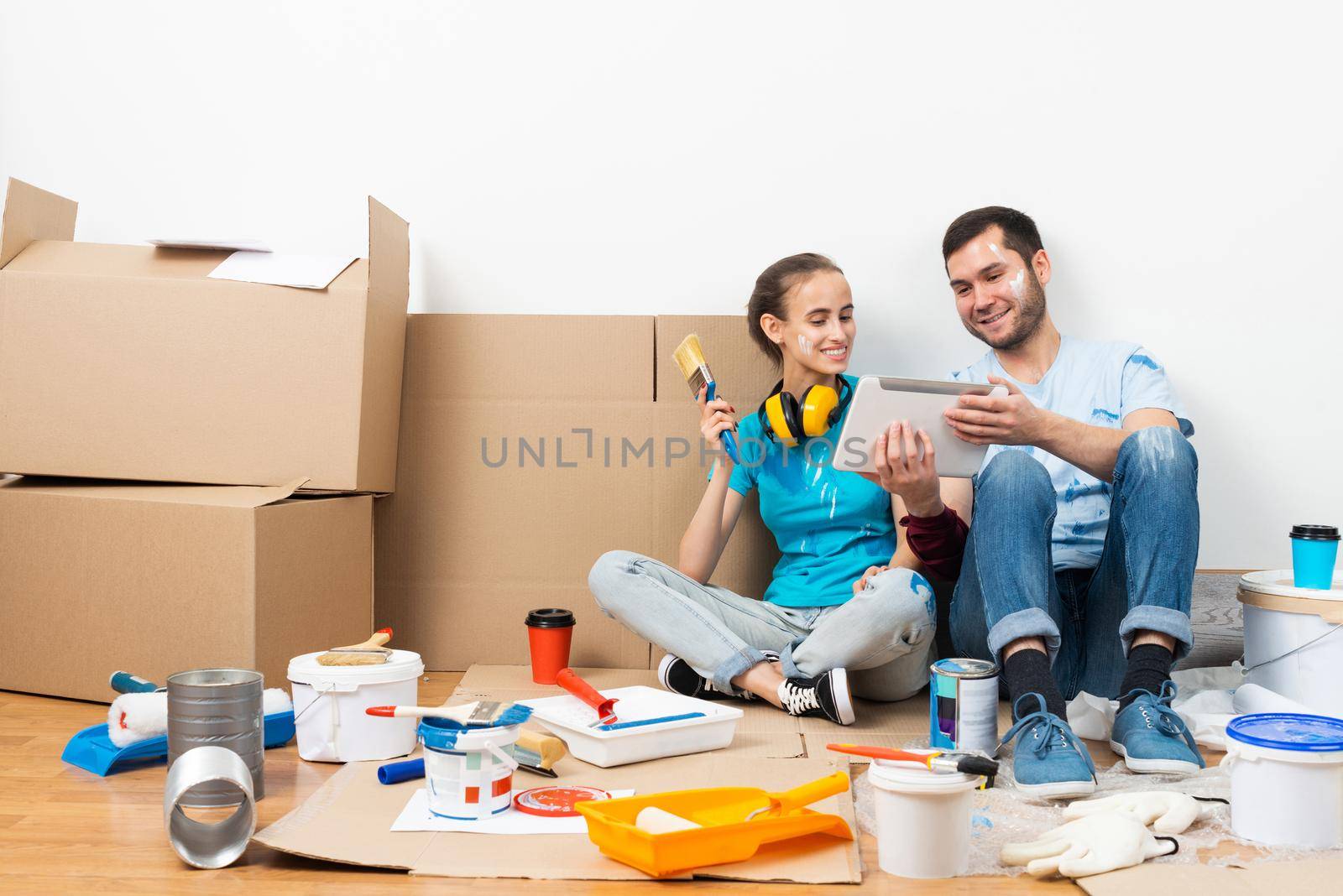 Happy couple sitting on floor and looking at tablet computer screen. Home remodeling and house interior redesign. Painting tools and materials on floor. Young family renovating their home after moving