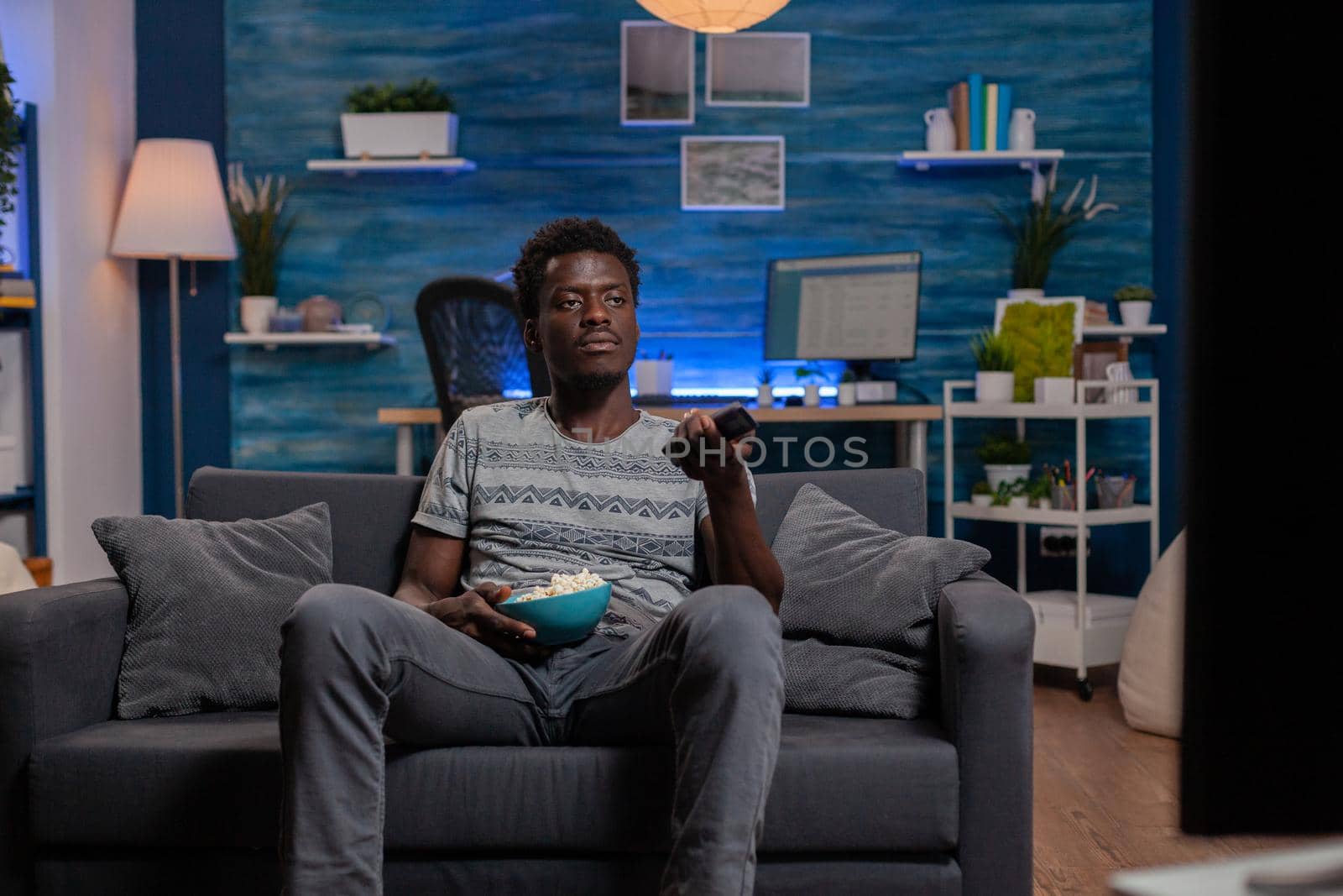 African american young man resting on sofa changing channel using remote by DCStudio