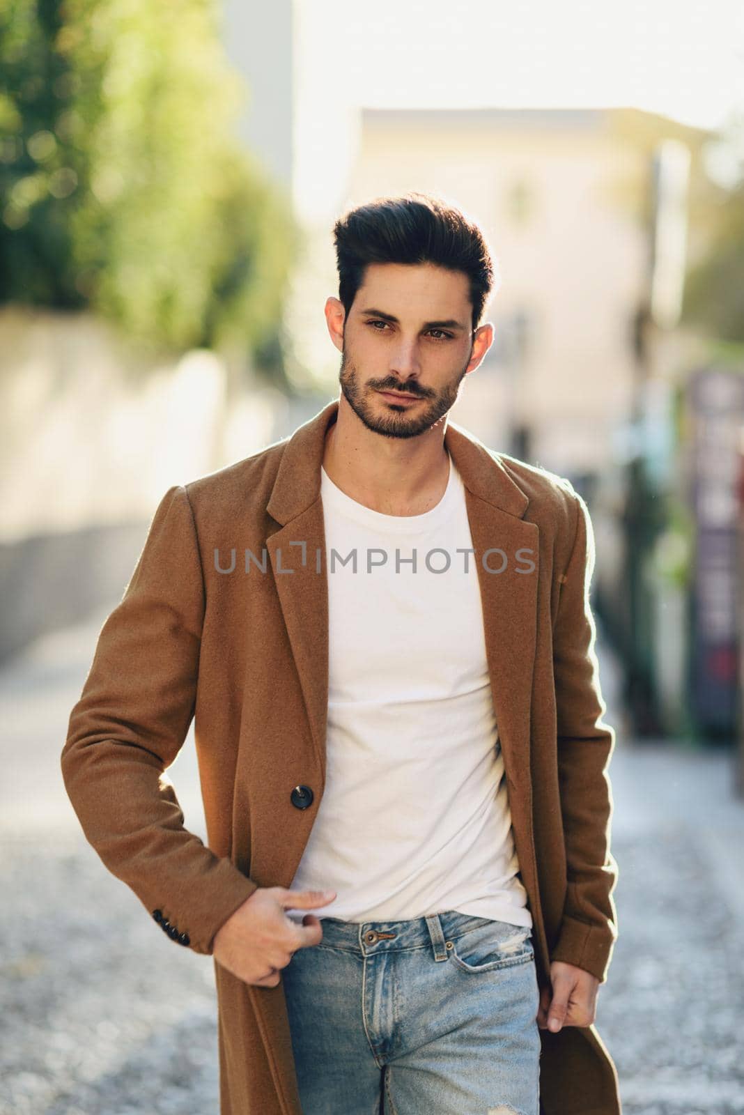 Young man wearing winter clothes in the street. Young bearded guy with modern hairstyle with coat, blue jeans and white t-shirt.