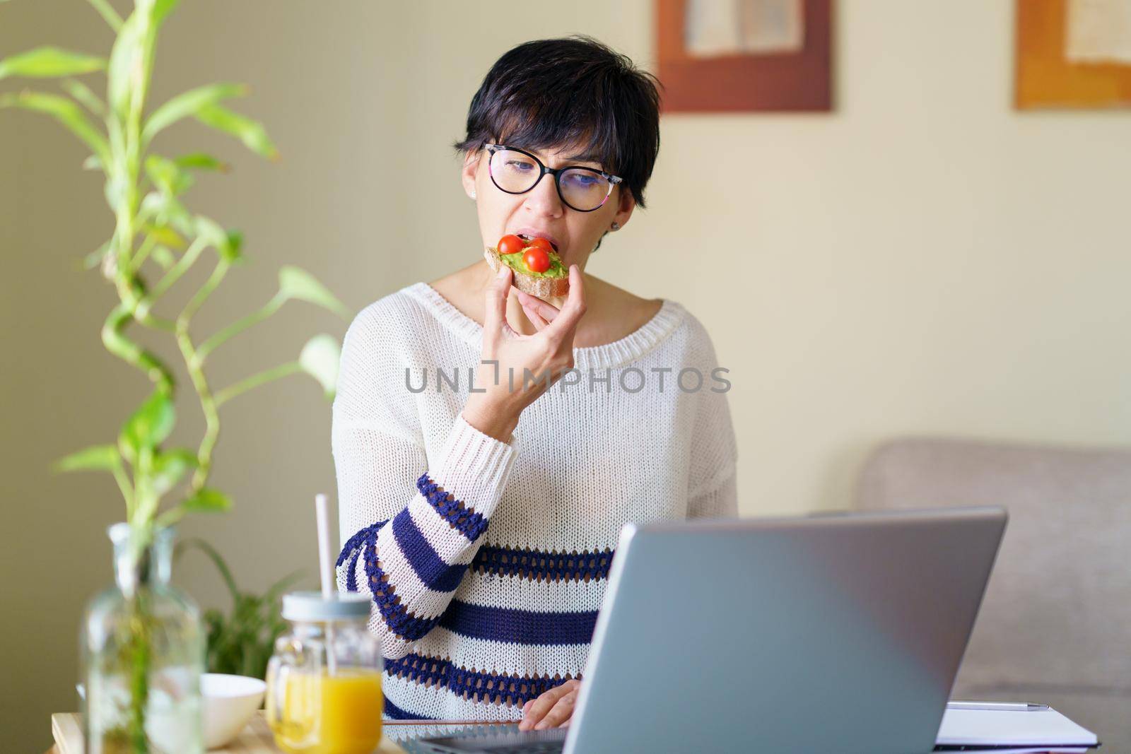 Woman eating some healthy food, while teleworking from home on her laptop. Female in her 50s