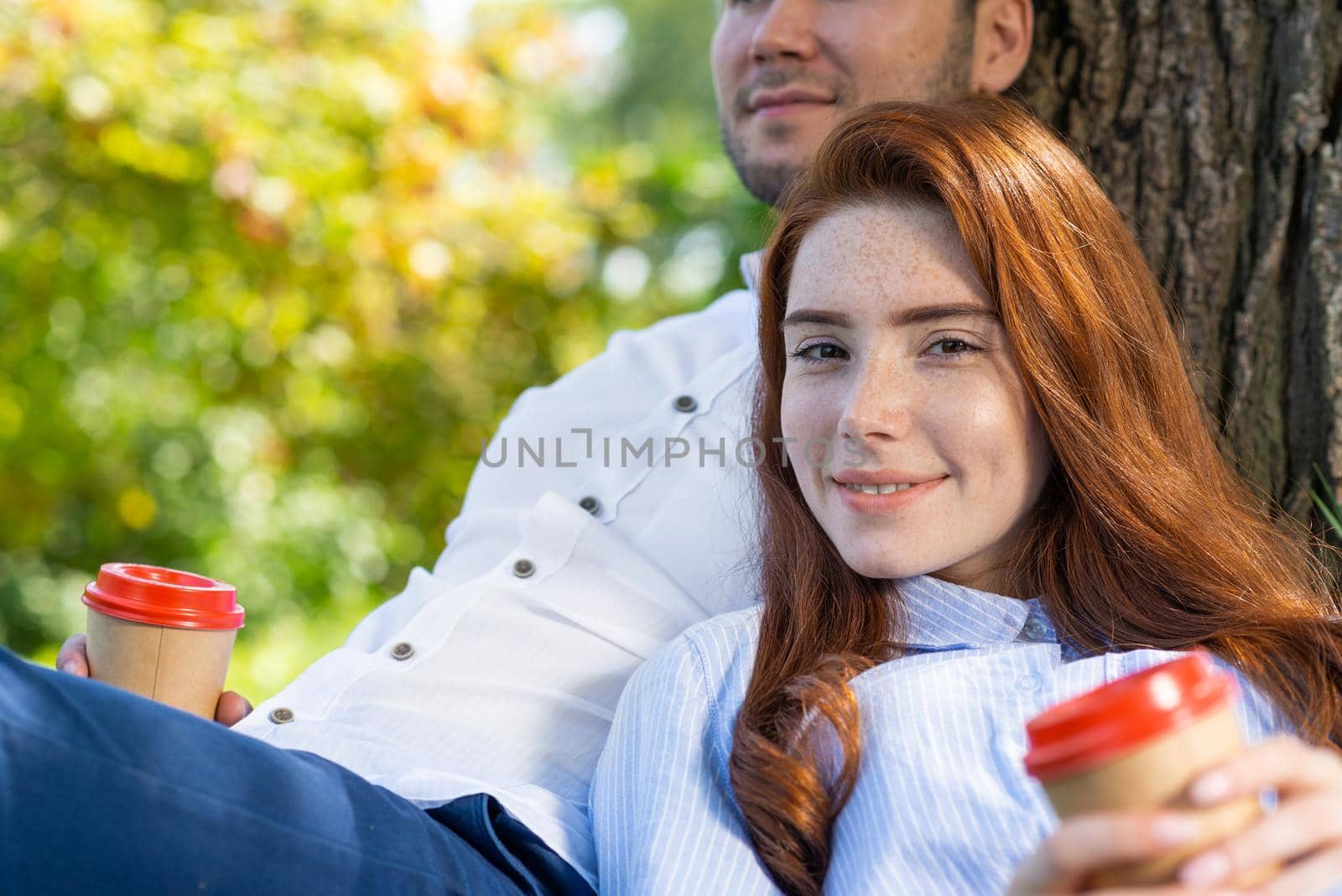 Young couple relaxing with coffee under tree in park on sunny day. Happy couple in love spend time outdoors together. Handsome man and pretty redhead girl sitting on green grass leaning against tree