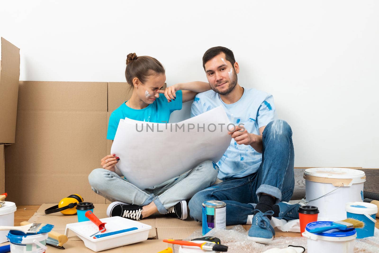 Young man and woman together planning their home renovation. Cardboard boxes, painting tools and materials on floor. House remodeling and interior renovation. People looking at blueprint at home.