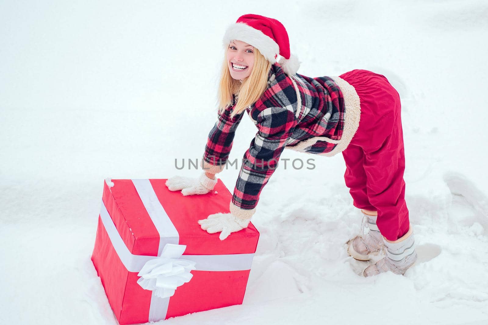 Winter woman with gift. Woman in Santa Claus dress giving you big red gift over white snow background. Happy woman standing on white snow background and holding a giant red gift box with both hands