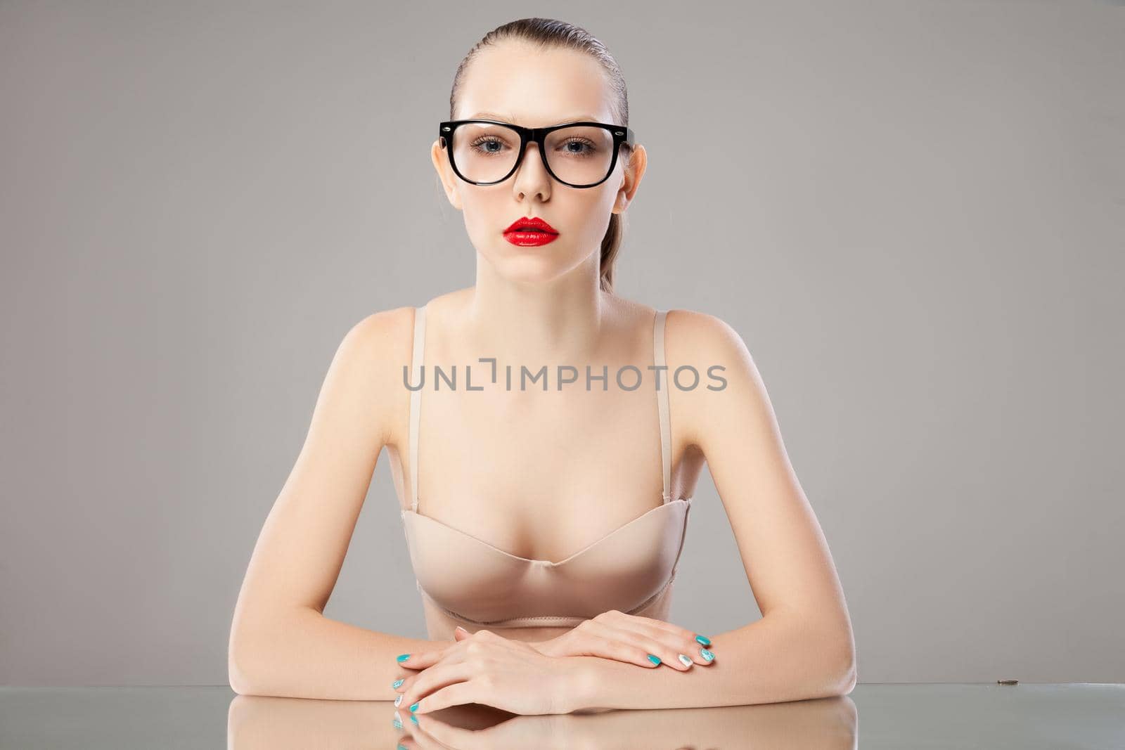 Young woman in bra and glasses with red lips looking at camera while sitting at table