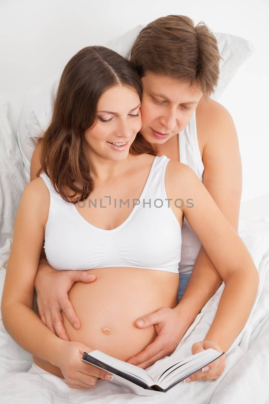 Close-up of a delighted pregnant woman and her husband reading book in the bed at home