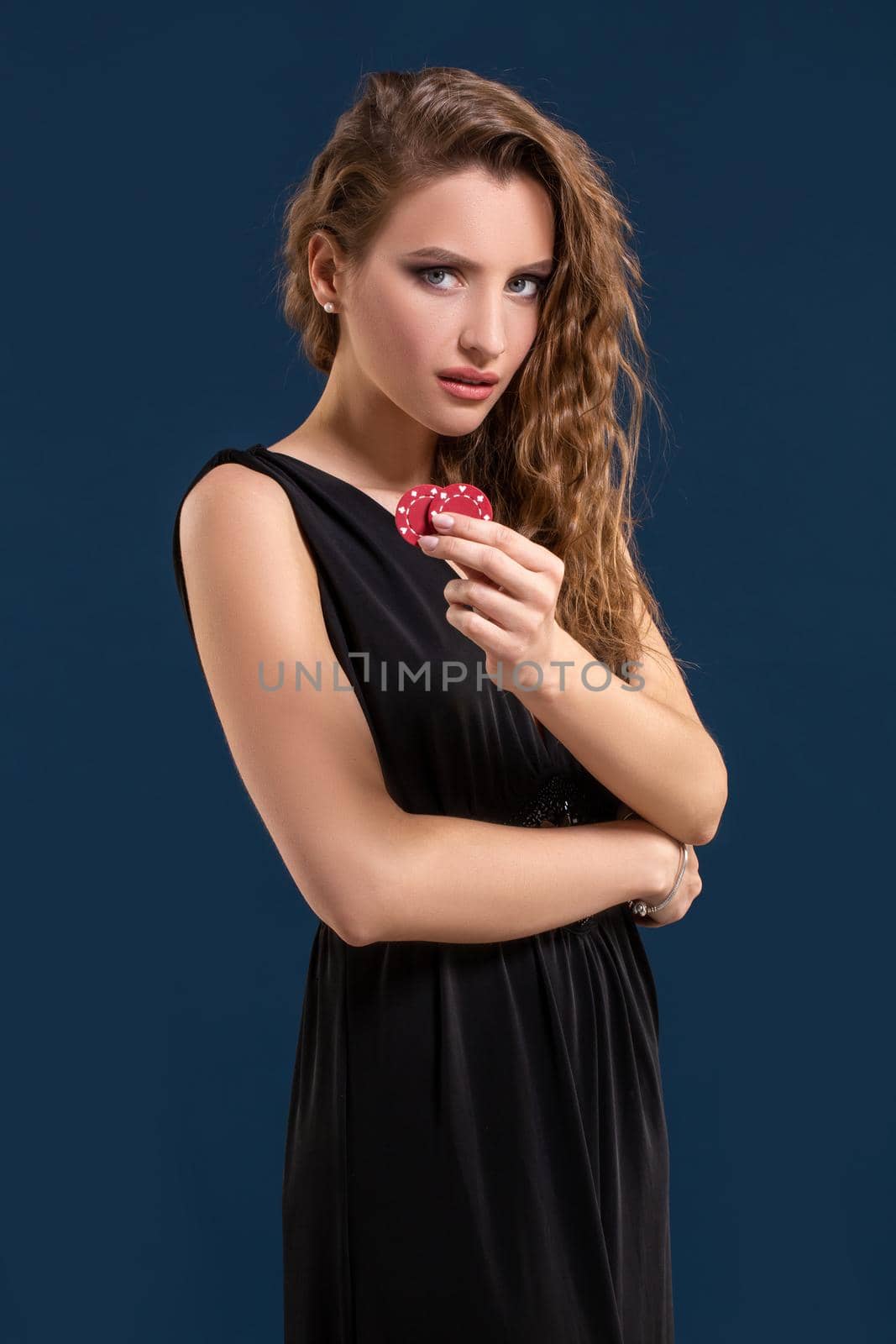Portrait of the female gambler at the casino keeping poker chips in the hand on dark blue background. Beautiful woman in black dress holds poker chips