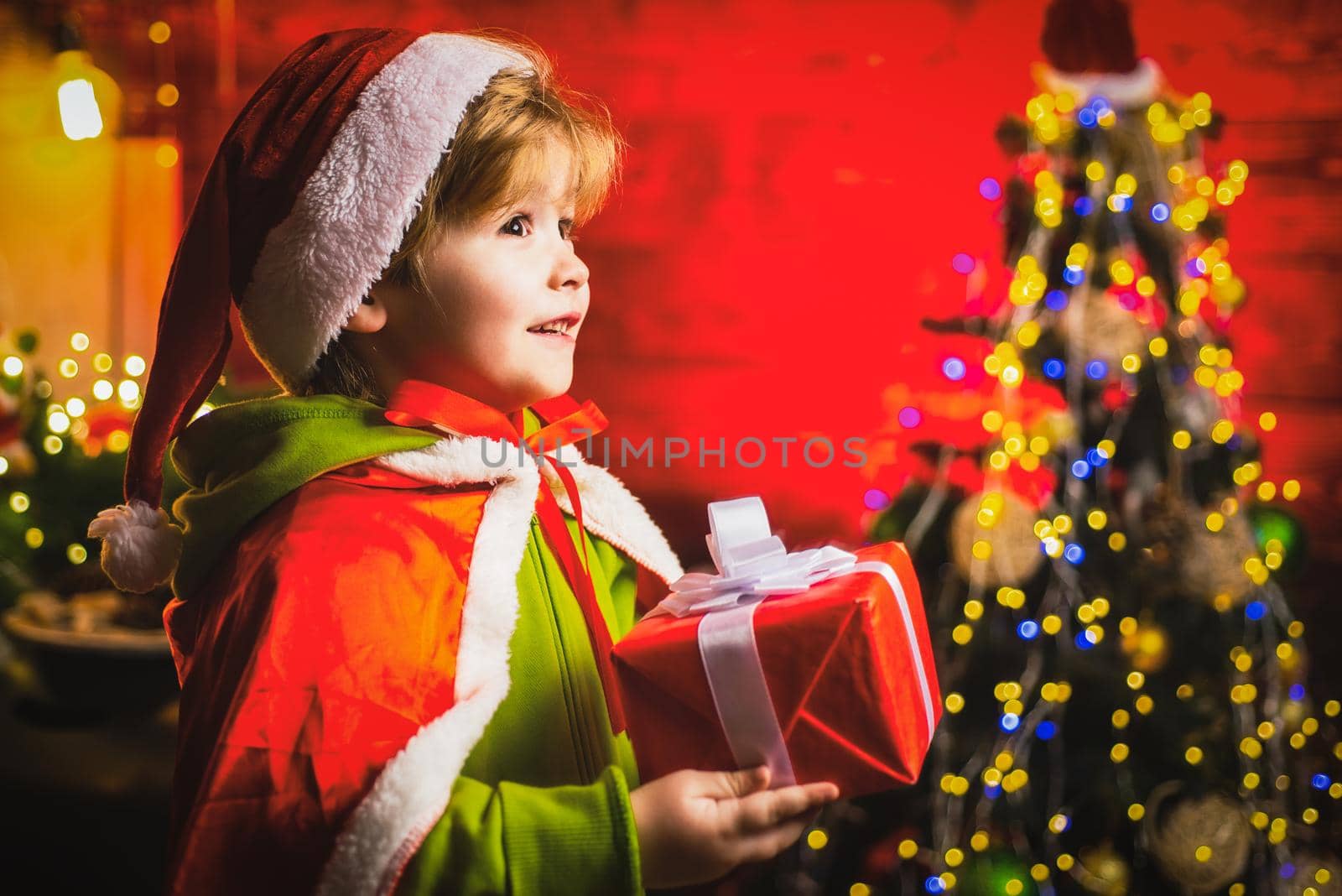 Happy little boy by near Christmas tree with Christmas gift. Little kid is wearing Santa clothes. Christmas kids concept. by Tverdokhlib