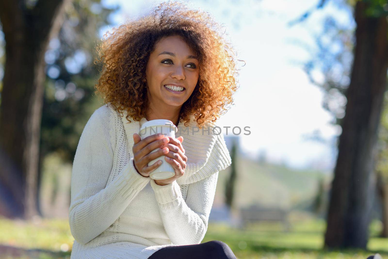 Beautiful young African American woman with afro hairstyle and green eyes wearing white winter dress. Girl drinking coffee in park sitting on grass wearing casual clothes smiling.