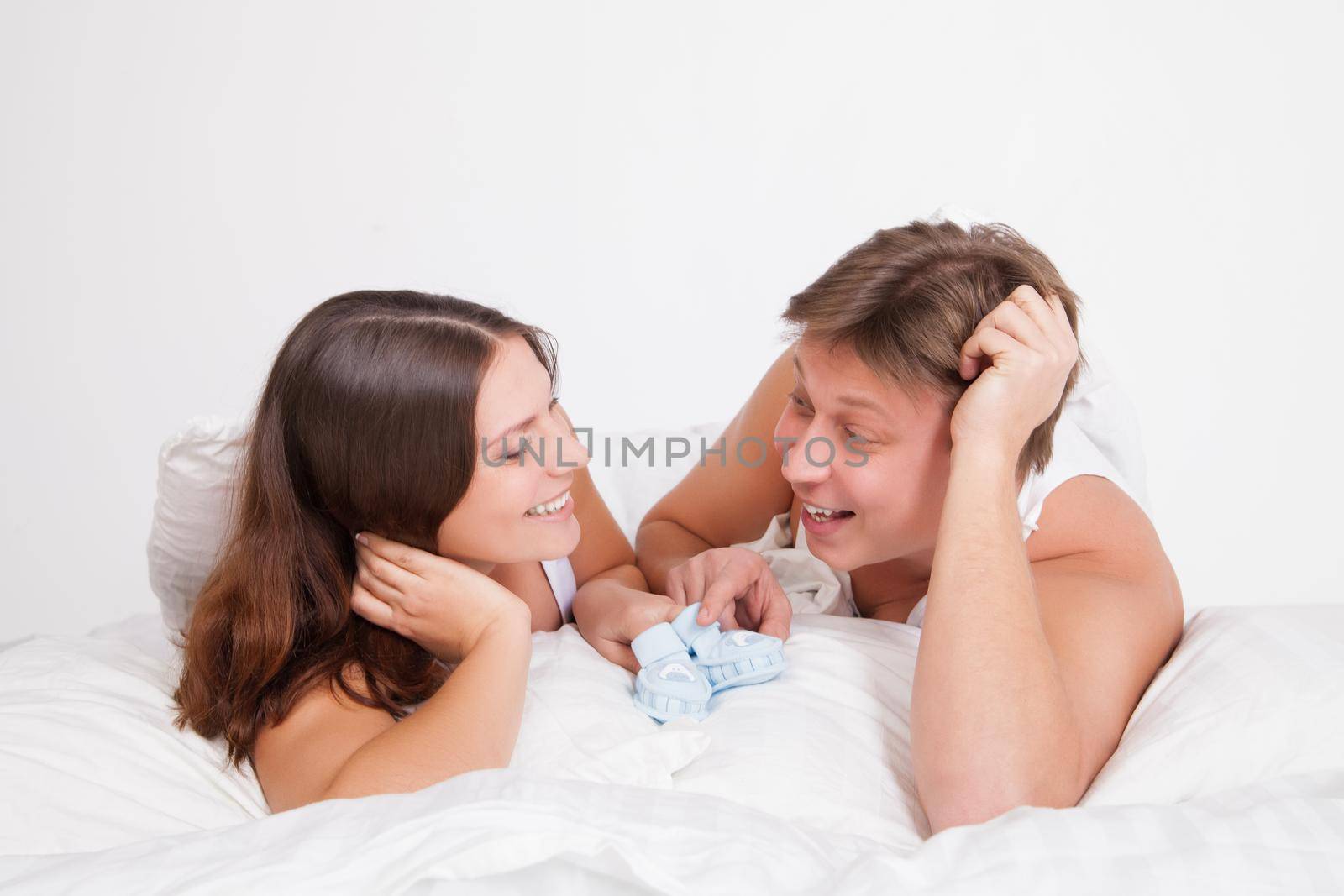 happy young pregnant woman with husband on white bed in light bedroom with baby booties