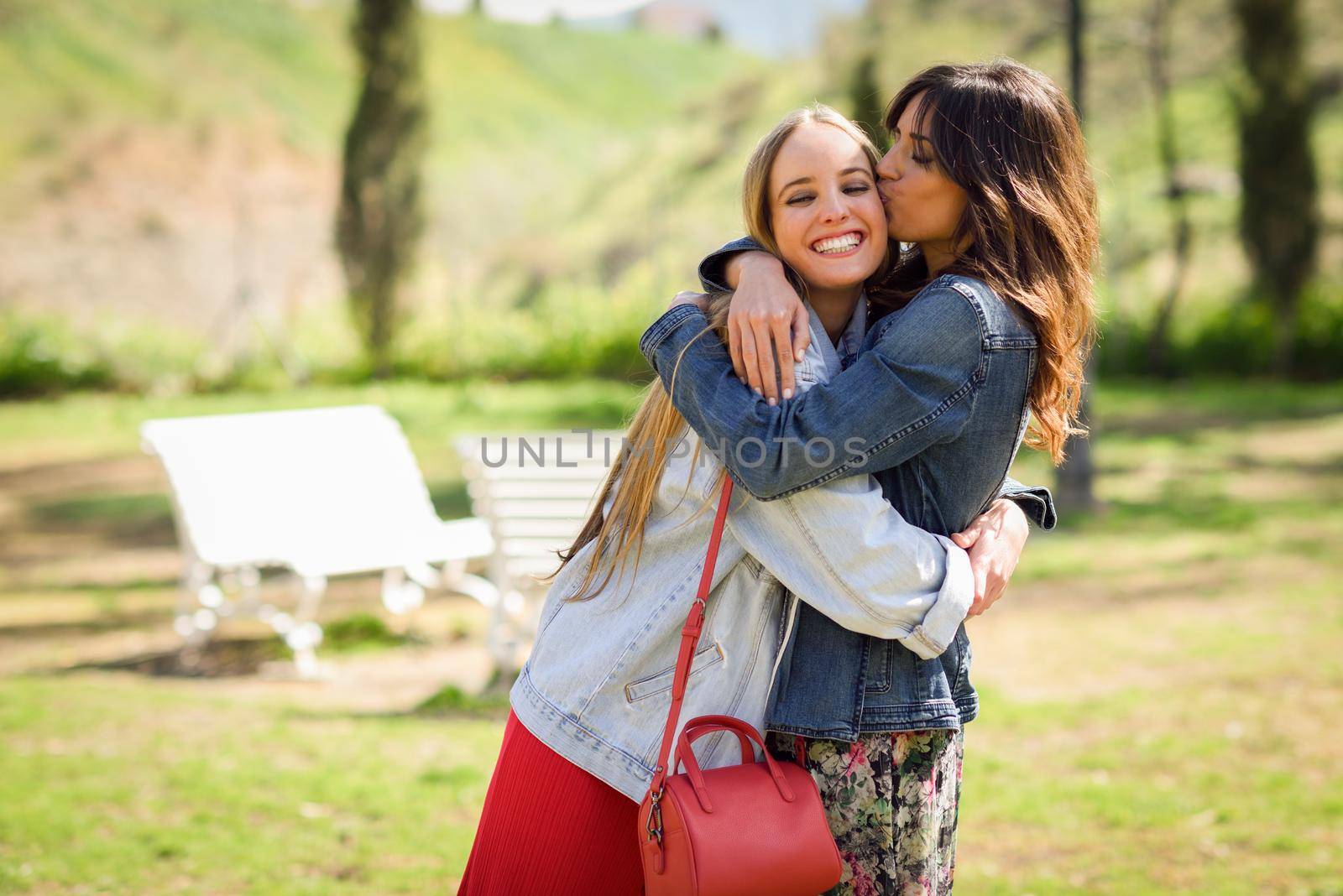 Young woman kissing her friend face outdoors. Blonde and brunette girls wearing casual clothes outdoors.